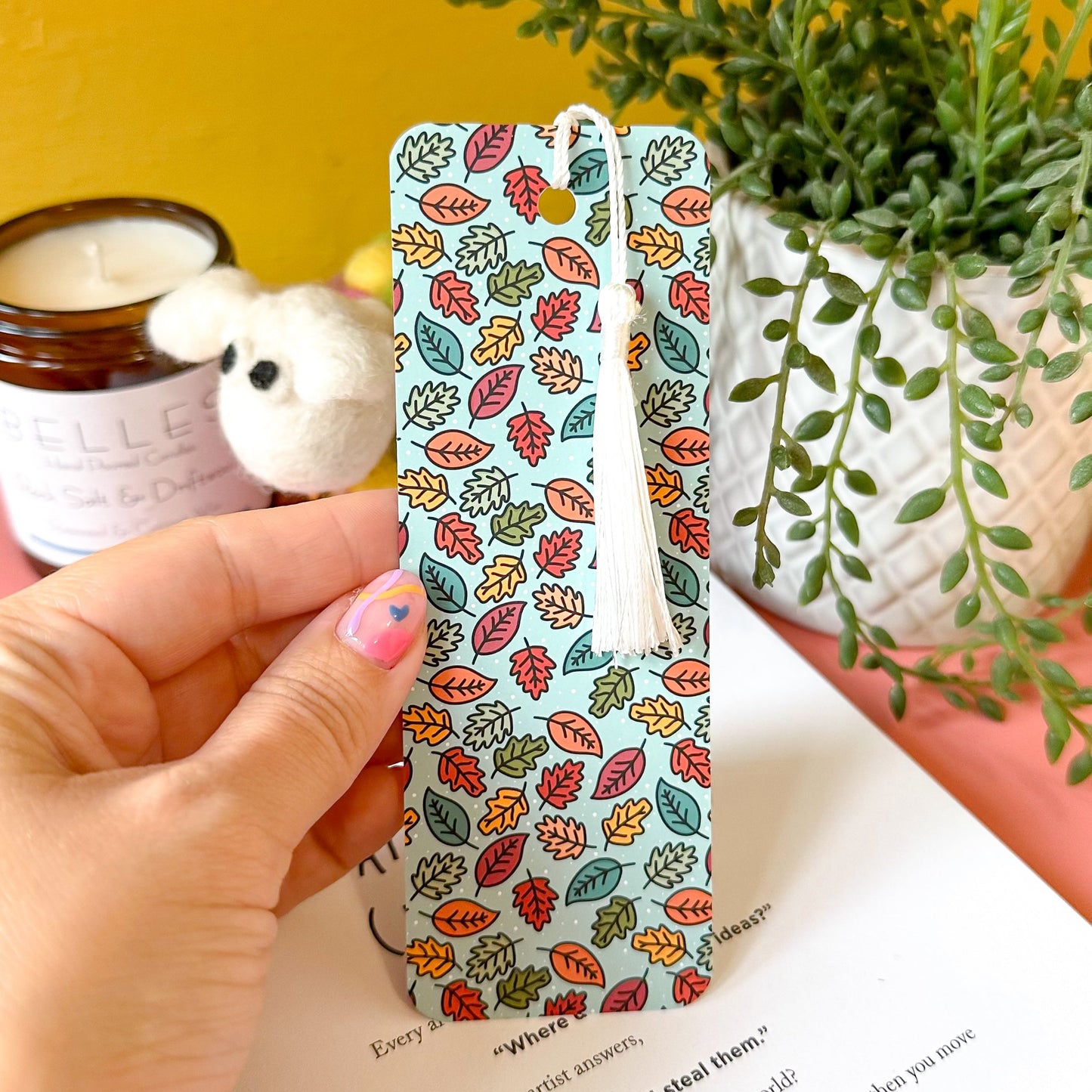 bookmark with leaf pattern held by a hand, with a plant and candle in the background