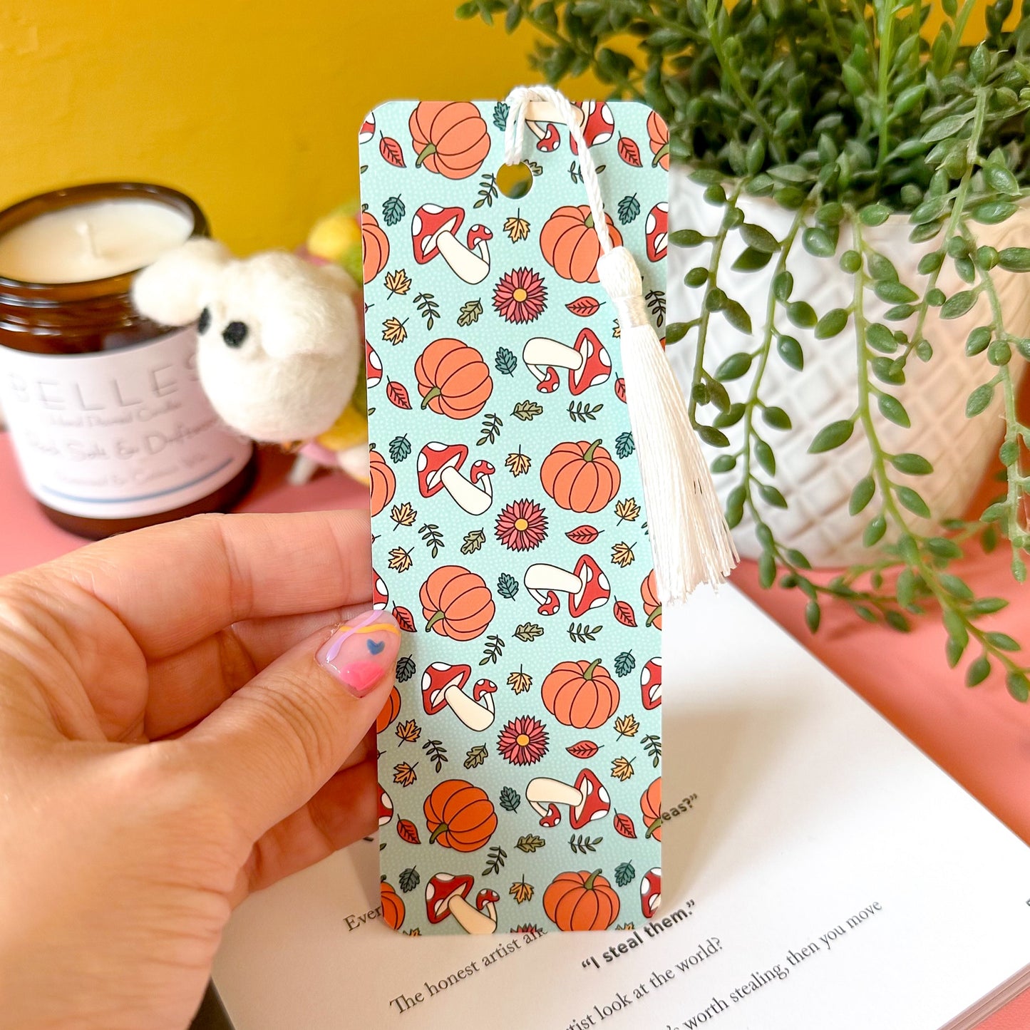 Hand holding a patterned bookmark with pumpkins and mushrooms on a book with a yellow background.