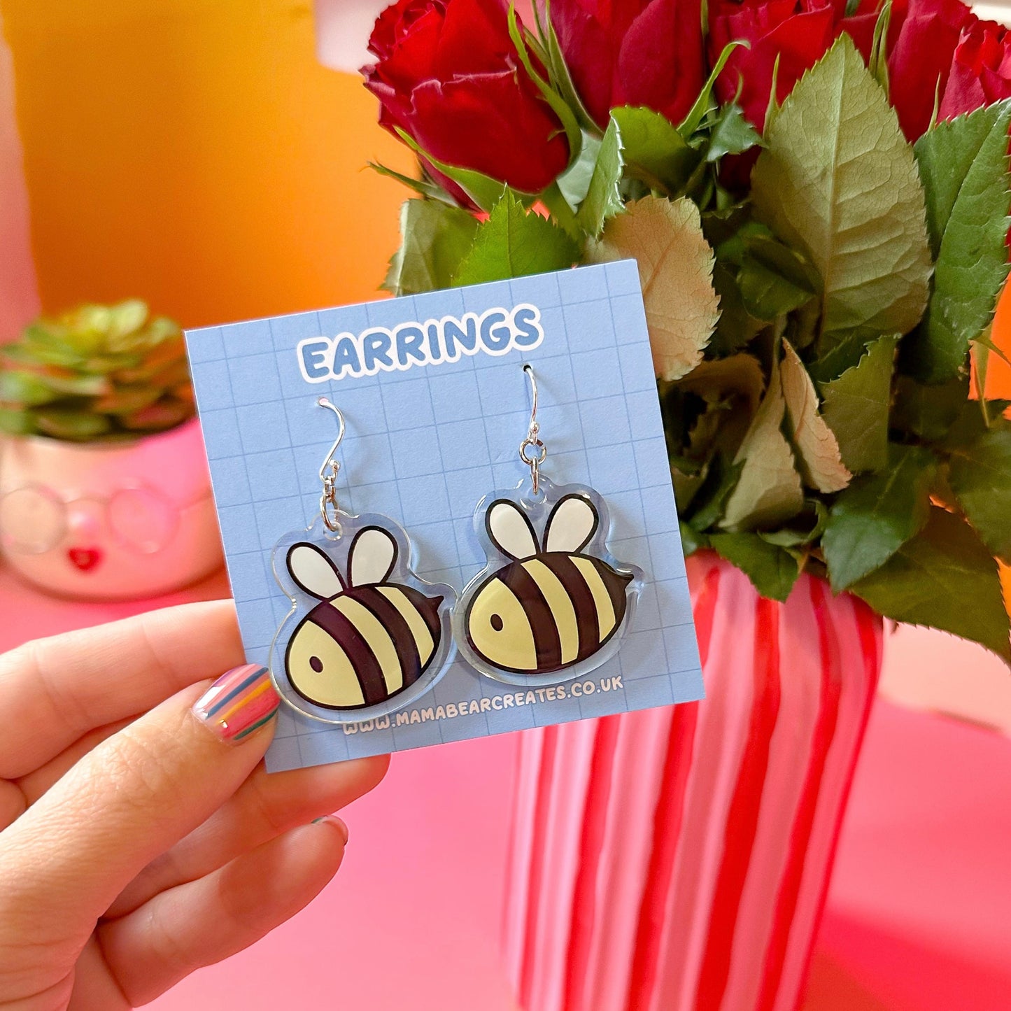 A pair of bee-shaped earrings on a blue earring card, displayed in front of a bouquet of roses.