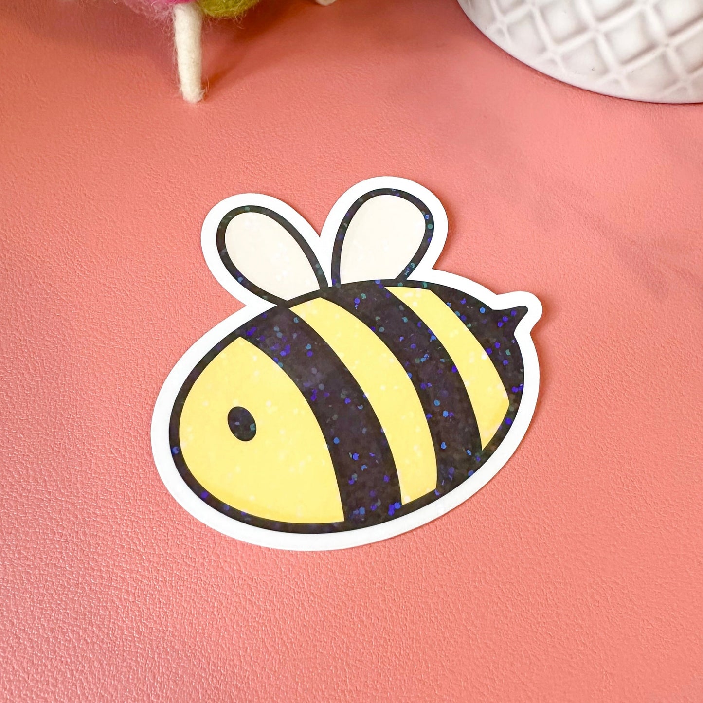 Sticker of a bee on a pink surface with a textured white object and colorful yarn in the background.