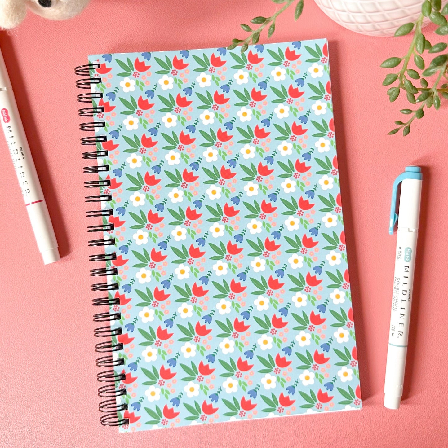 Notebook with floral pattern on a pink surface with pens and plants