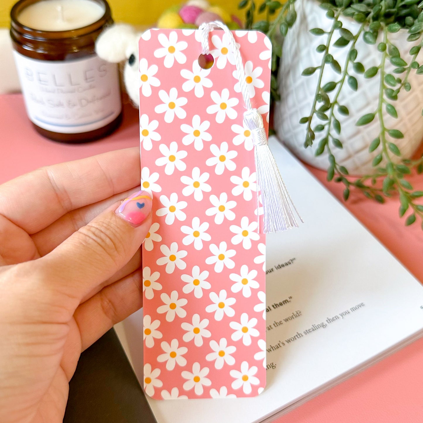 Hand holding a pink floral bookmark over an open book with a yellow wall and plant in the background.