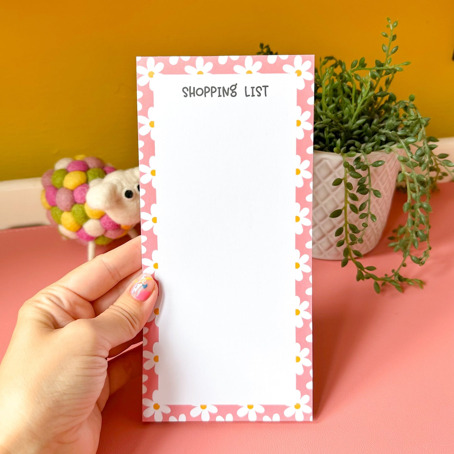 Hand holding a small notepad with 'Shopping List' text against a pink and yellow background.