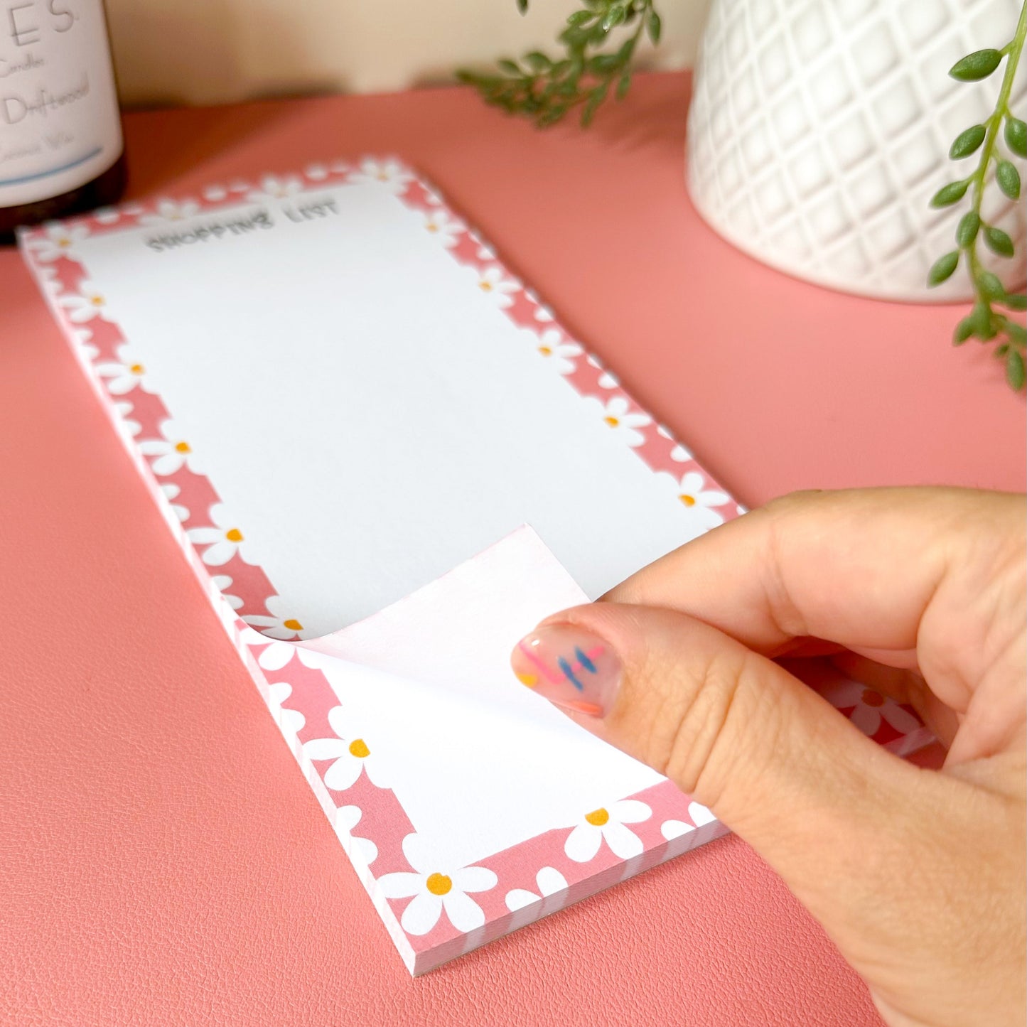 Hand holding a floral notepad on a pink surface with a plant in the background