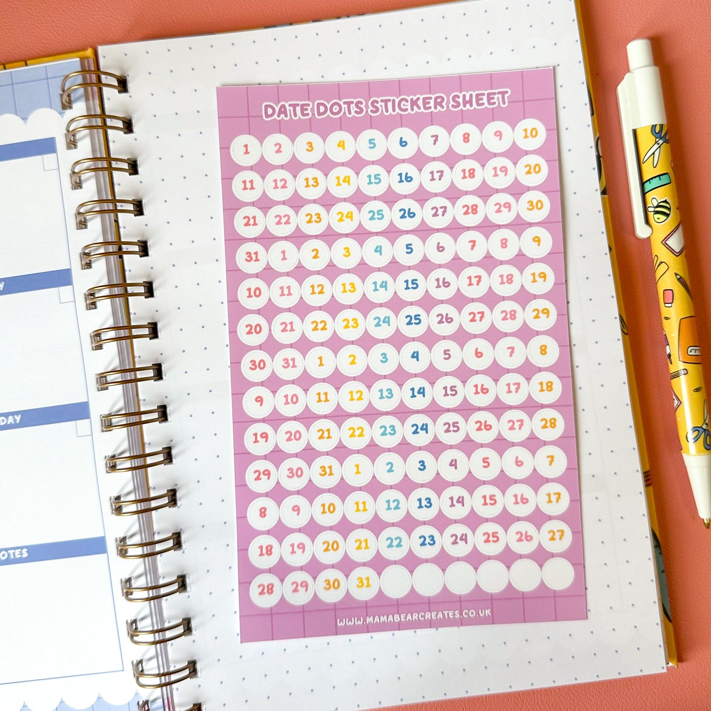 Date dot sticker sheet on a planner with a pen and plant on a pink background