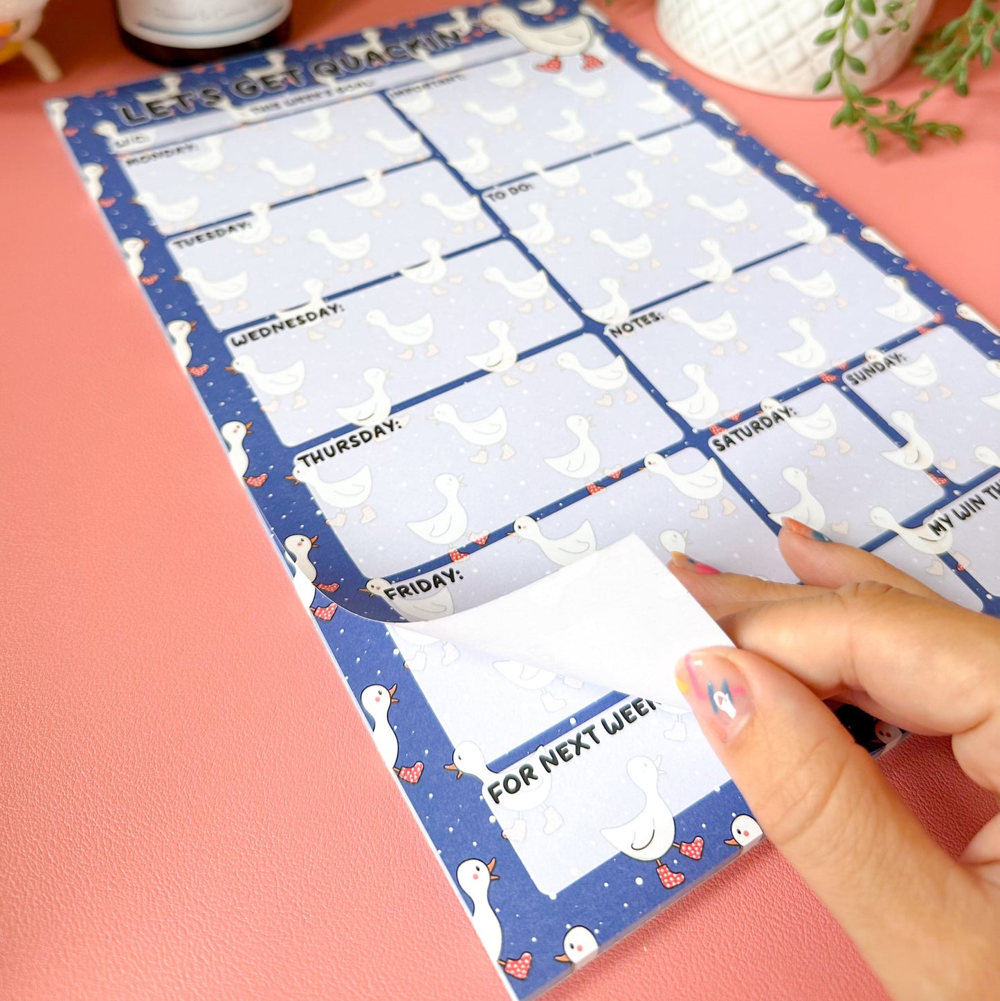 Weekly planner sheet with cartoon animals on a pink surface with a yellow wall and plant in the background.