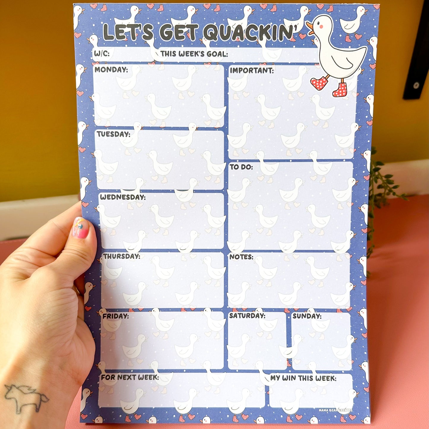 Weekly planner pad with a cartoon duck design held by a hand on a pink surface.