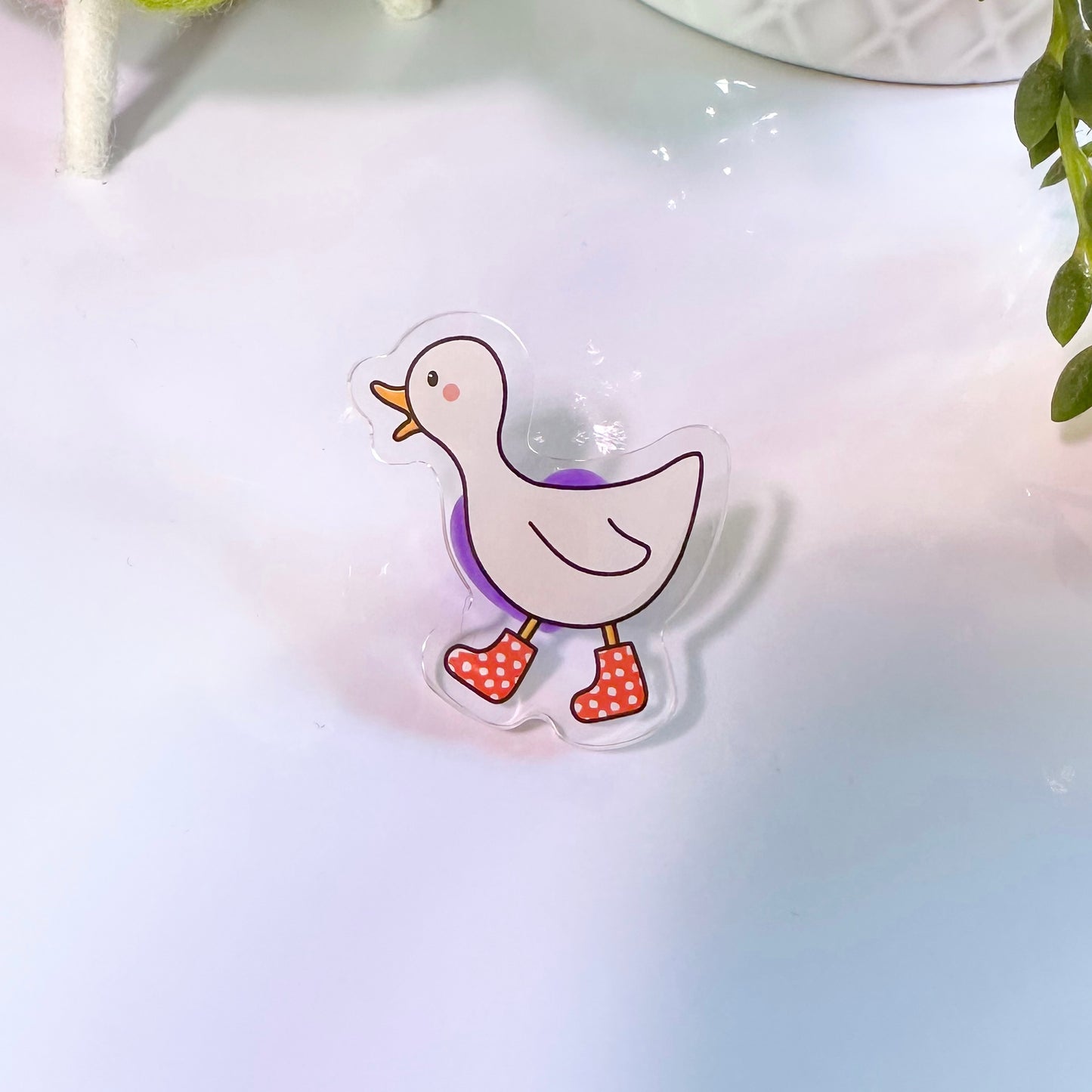 A pin badge featuring an acrylic depiction of a white duck with red boots, presented on a soft background.