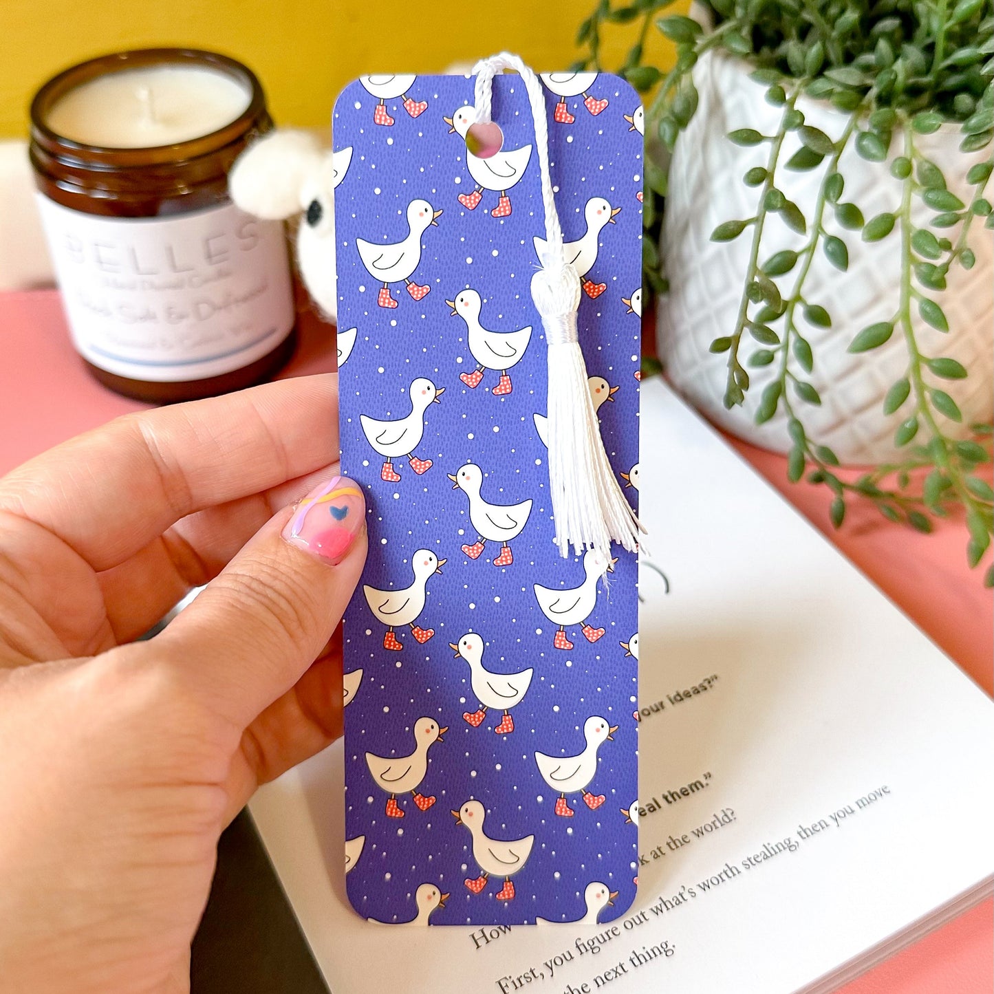 Hand holding a bookmark with duck pattern on a book with a candle and plant in the background