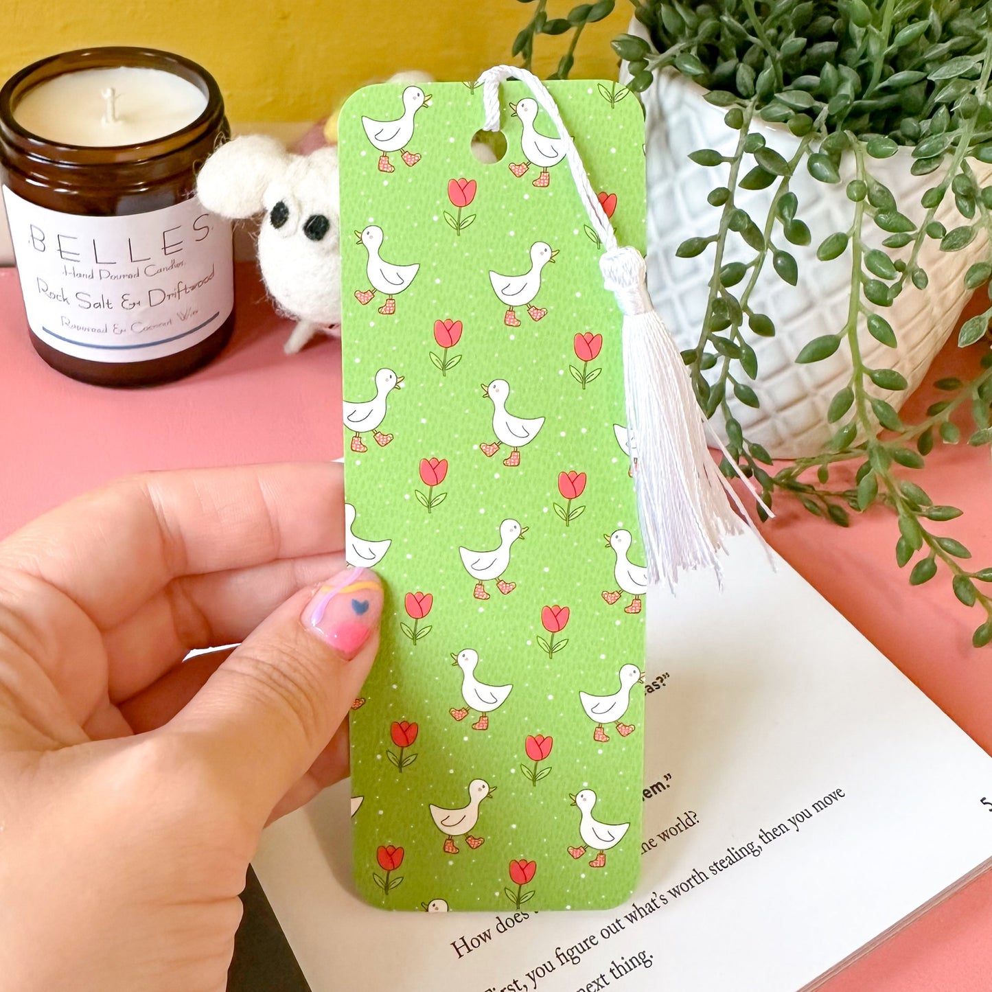 Hand holding a green bookmark with duck pattern on a book with a candle and plant in the background.