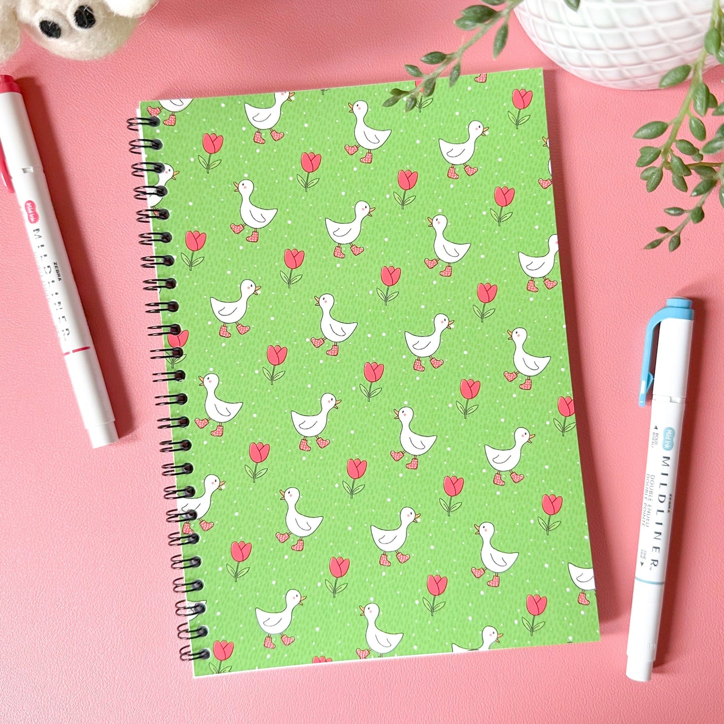 Green notebook with duck and flower pattern on a pink surface with pens and plants.