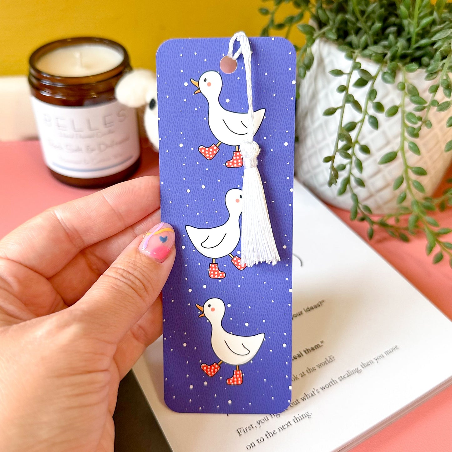 Hand holding a blue bookmark with bird illustrations on a book with a yellow background and plant.