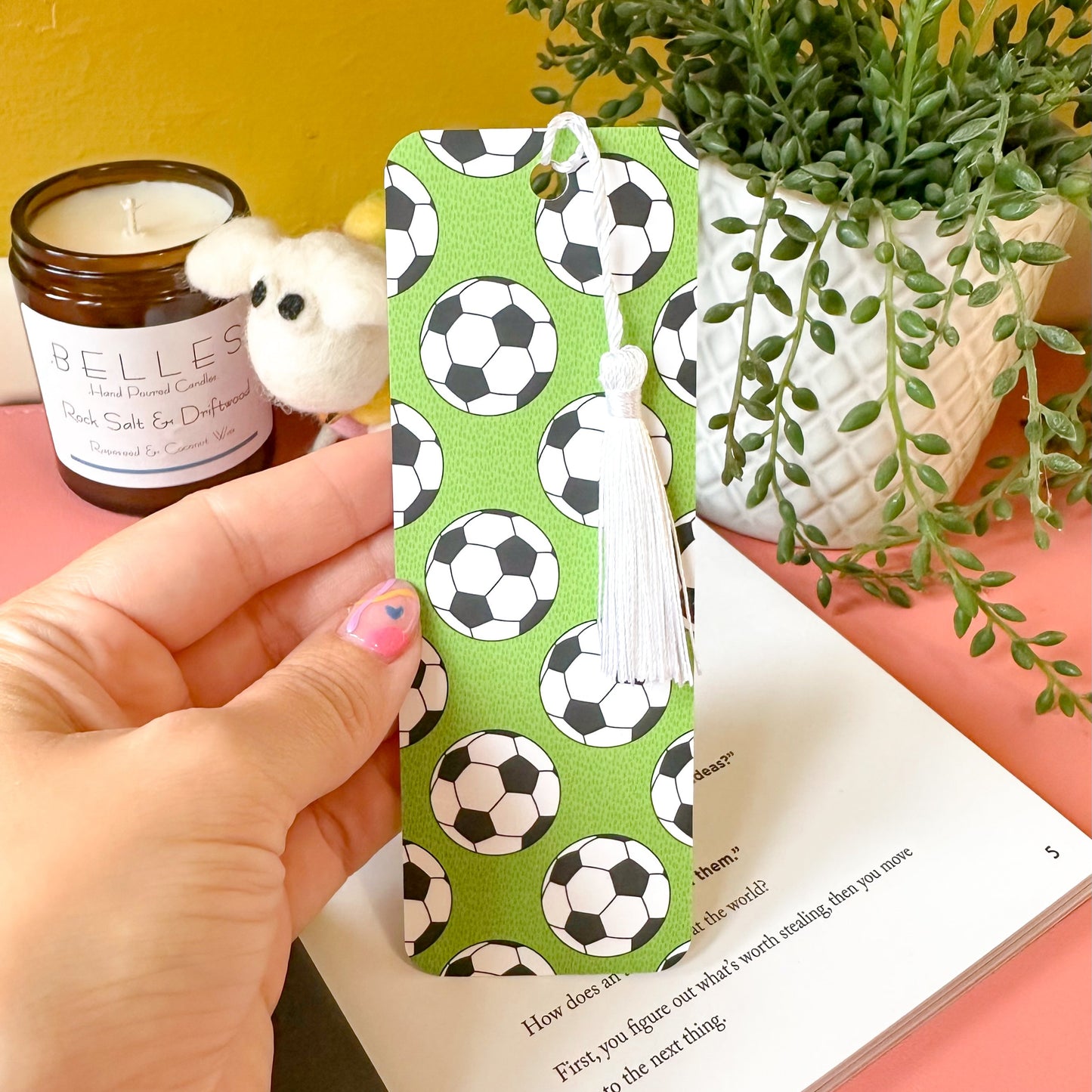 Hand holding a bookmark with football pattern in front of a book, candle, and plant.