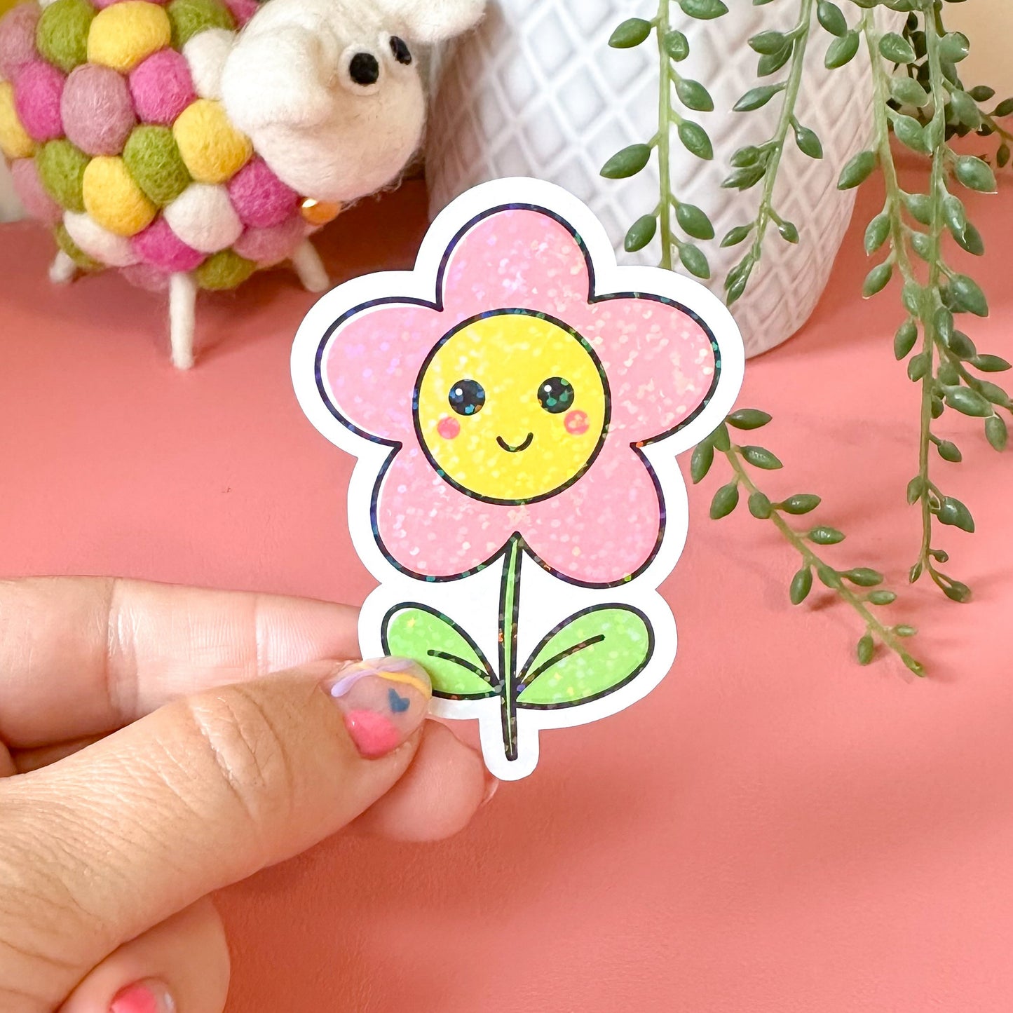 Hand holding a smiling flower sticker with a pink background featuring a sheep and plants.