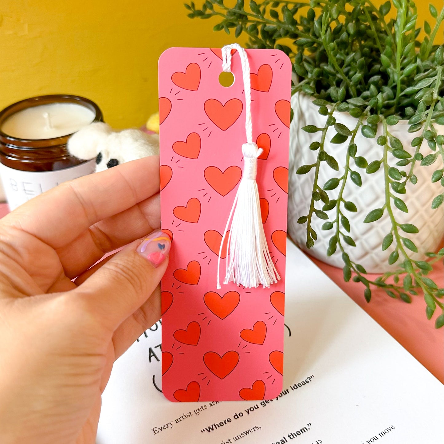 Pink bookmark with heart patterns held by a hand, with a plant and candle in the background.