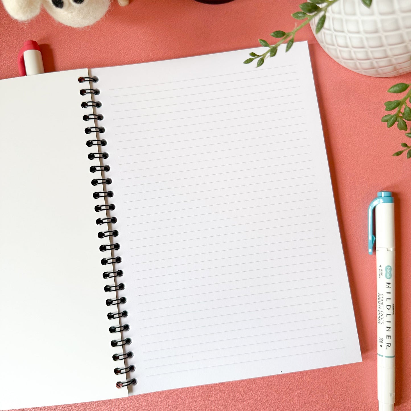 Open notebook with a pen on a pink surface with a plant and small bottle in the background