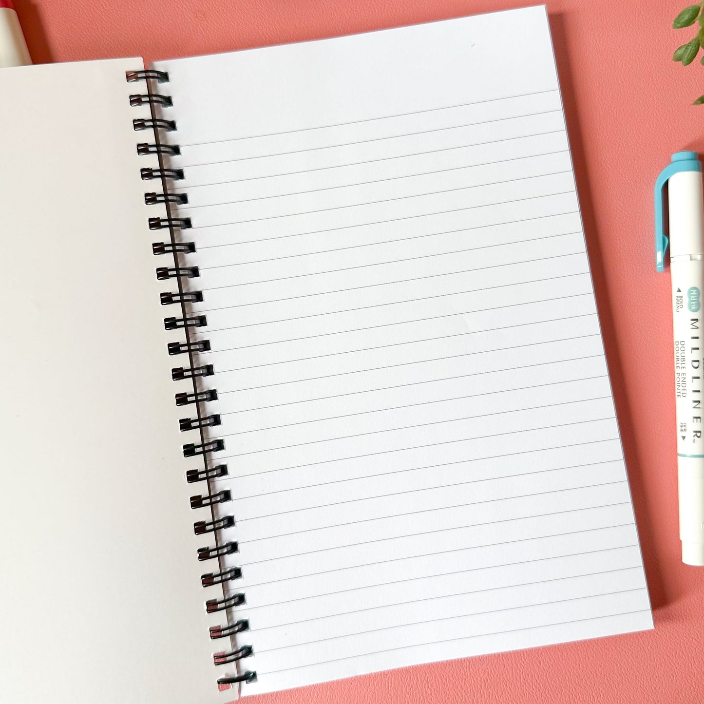 Open notebook with lined pages on a pink surface with a pen and decorative items.