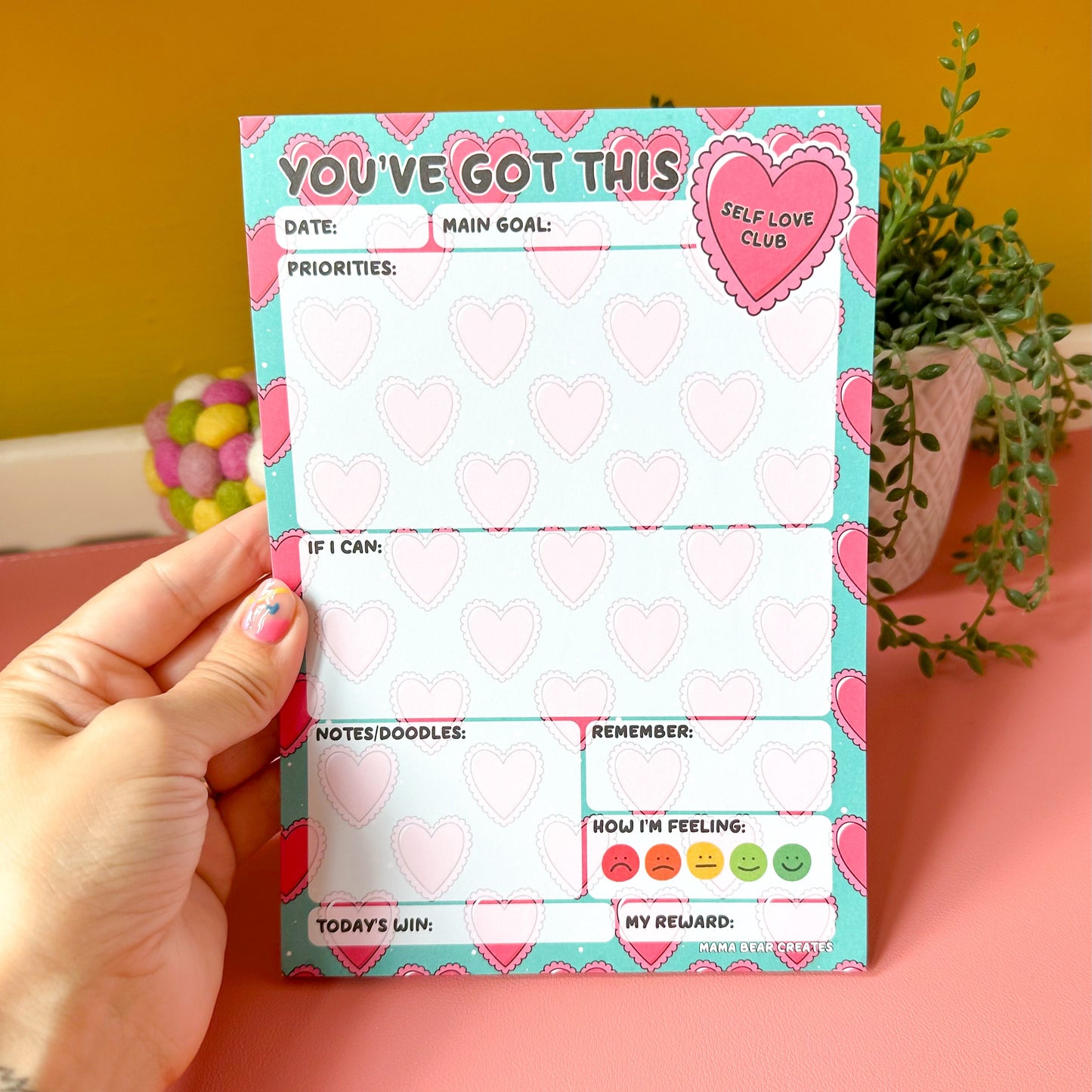 Hand holding a colorful notepad with heart designs against a pink background