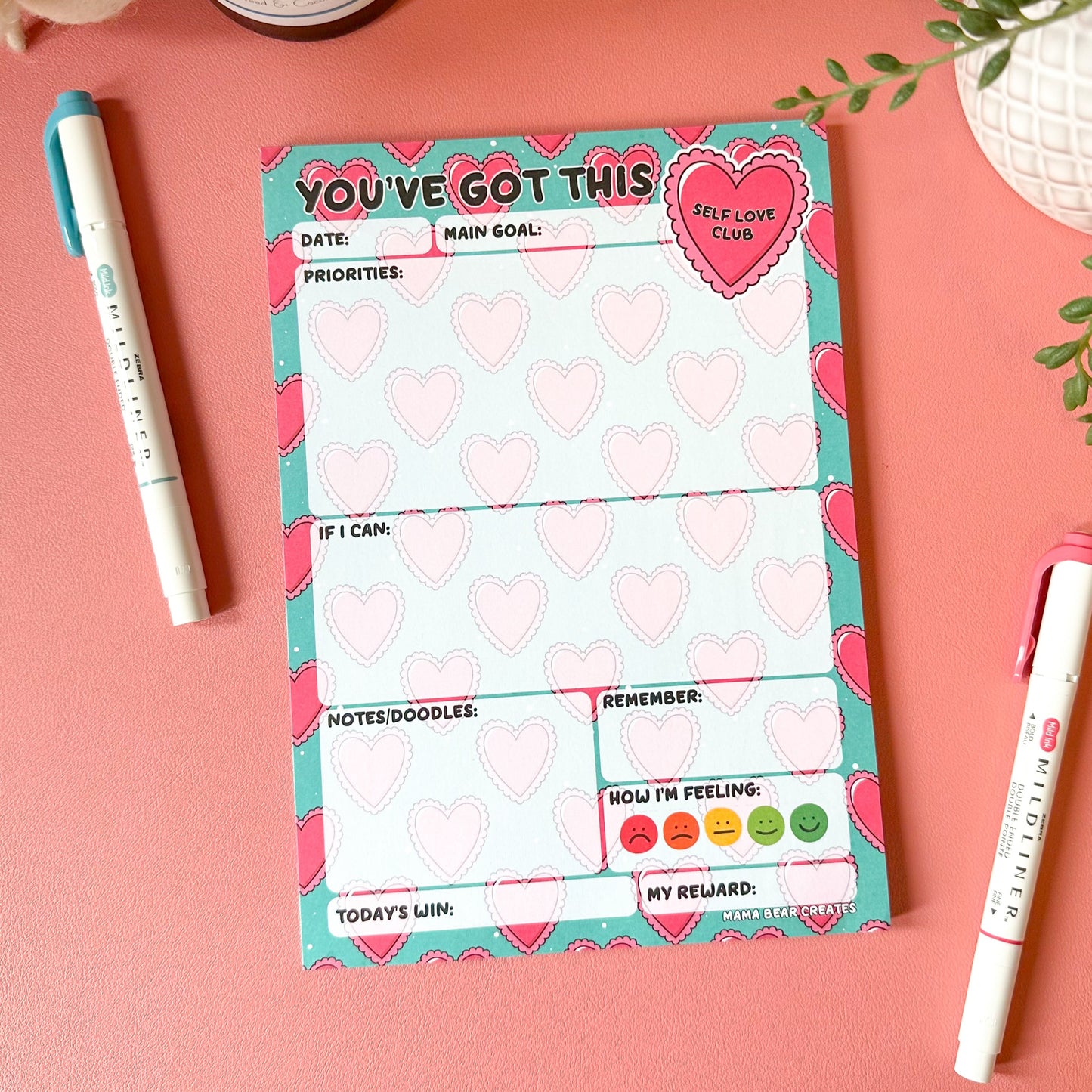 Motivational notepad with heart designs on a pink surface with pens and decor items.