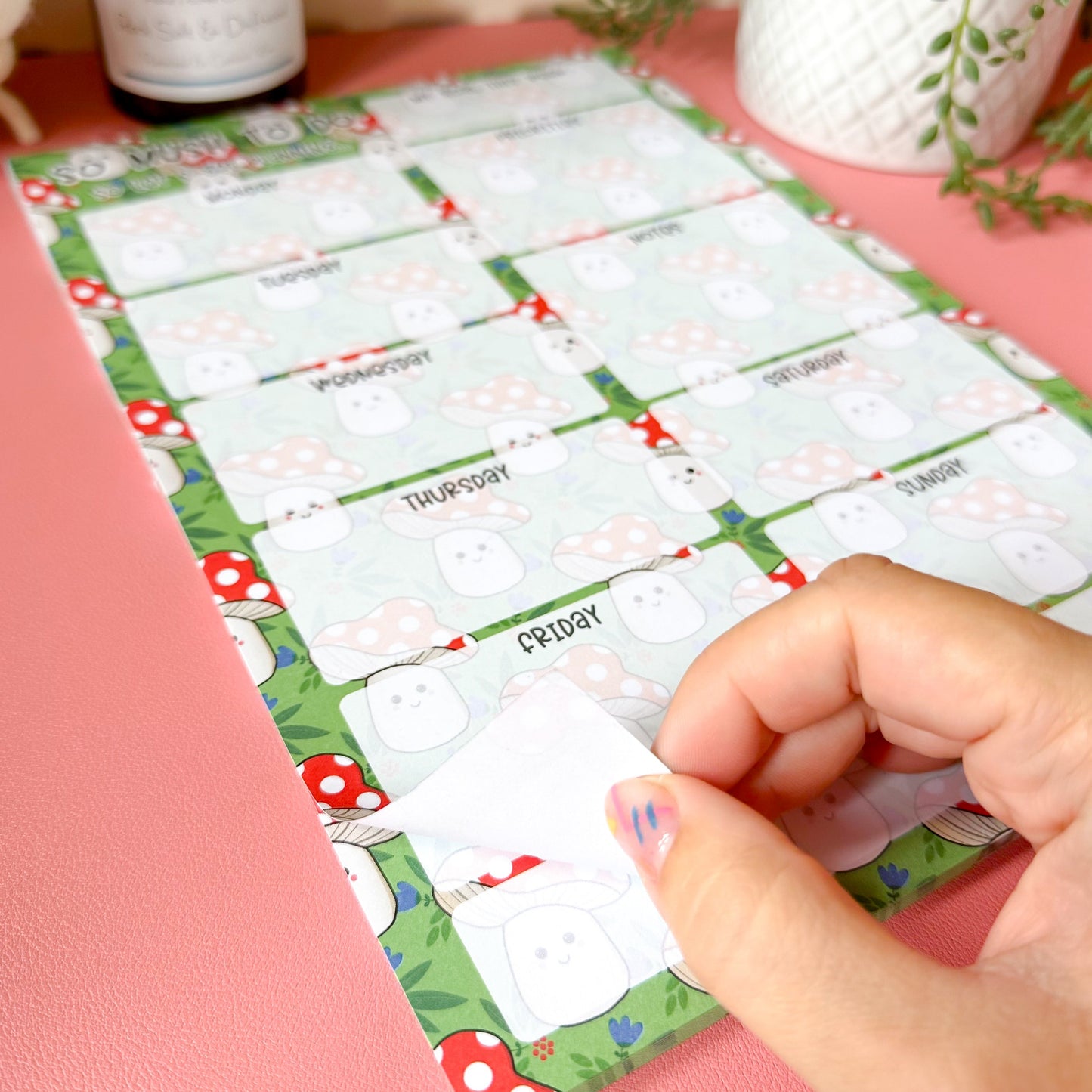 Sheet ofa4 planner pad holding one page, on a pink surface with a plant and candle in the background.