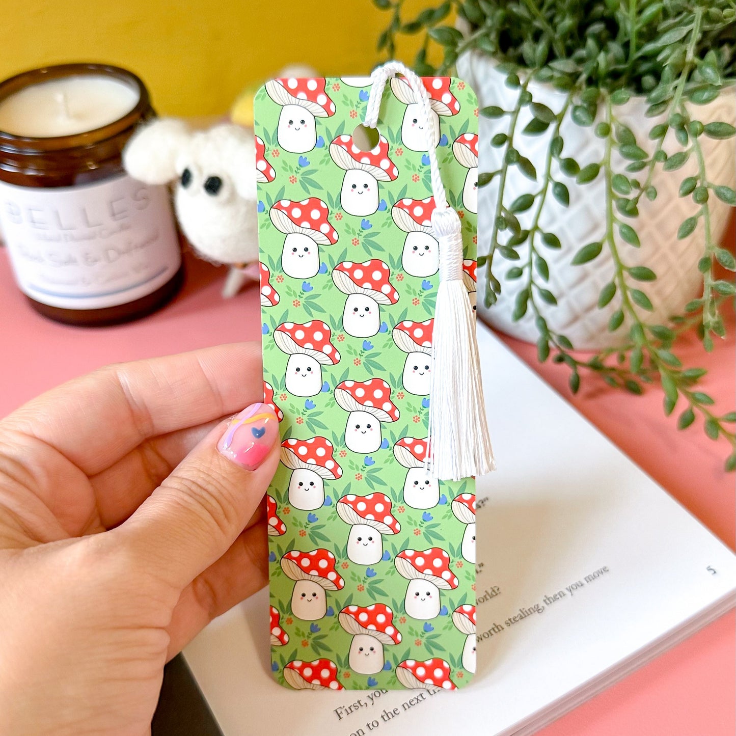 Hand holding a mushroom patterned bookmark with a yellow wall, candle, and plant in the background