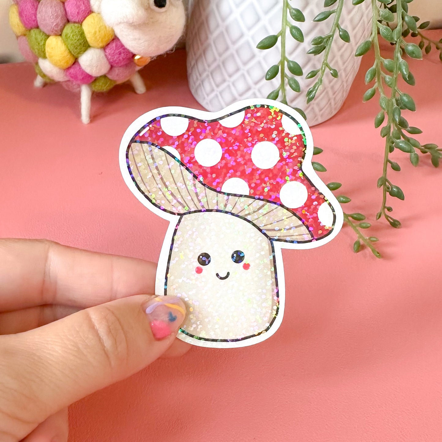 Hand holding a cute cartoon mushroom sticker with a pink polka dot cap against a pink background.