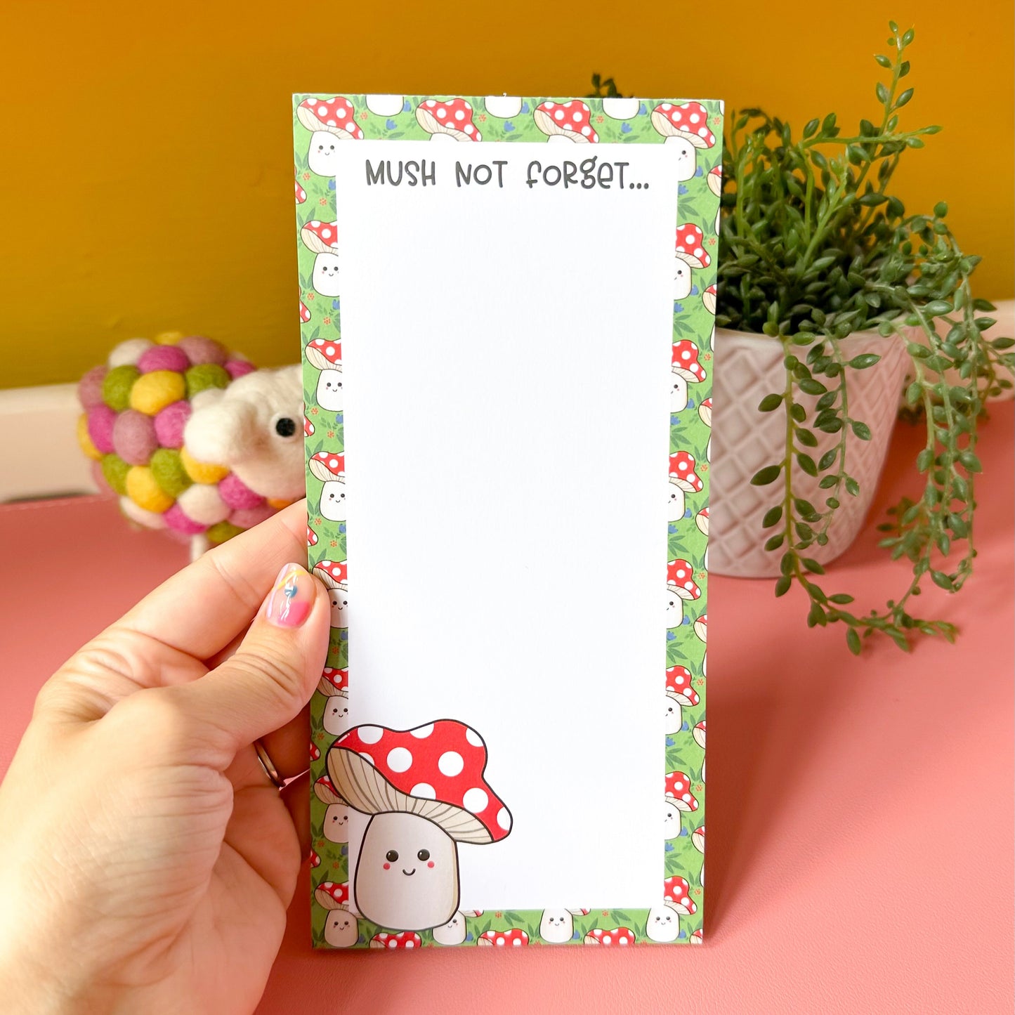 Notepad with a cute mushroom design held by a hand against a pink and yellow background.