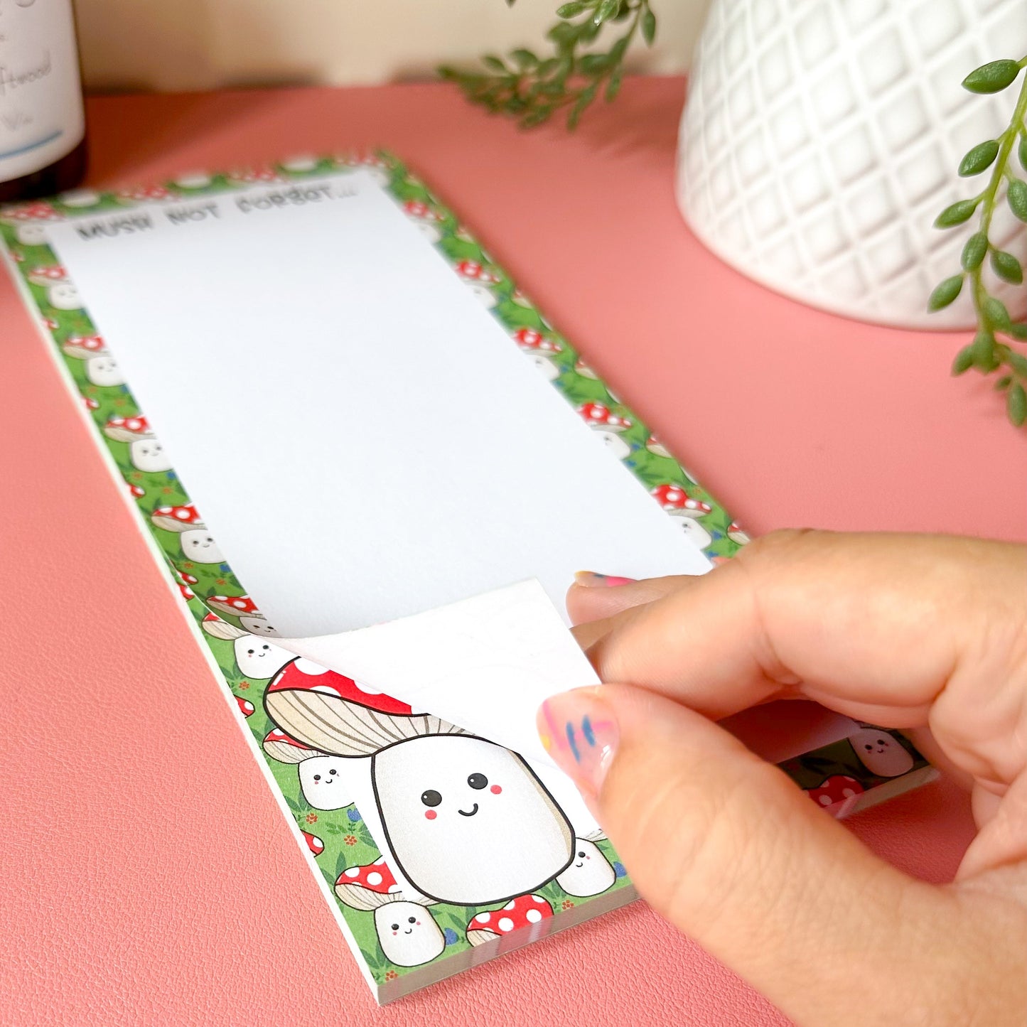 Hand holding a small notepad with a cute mushi design on a pink surface.