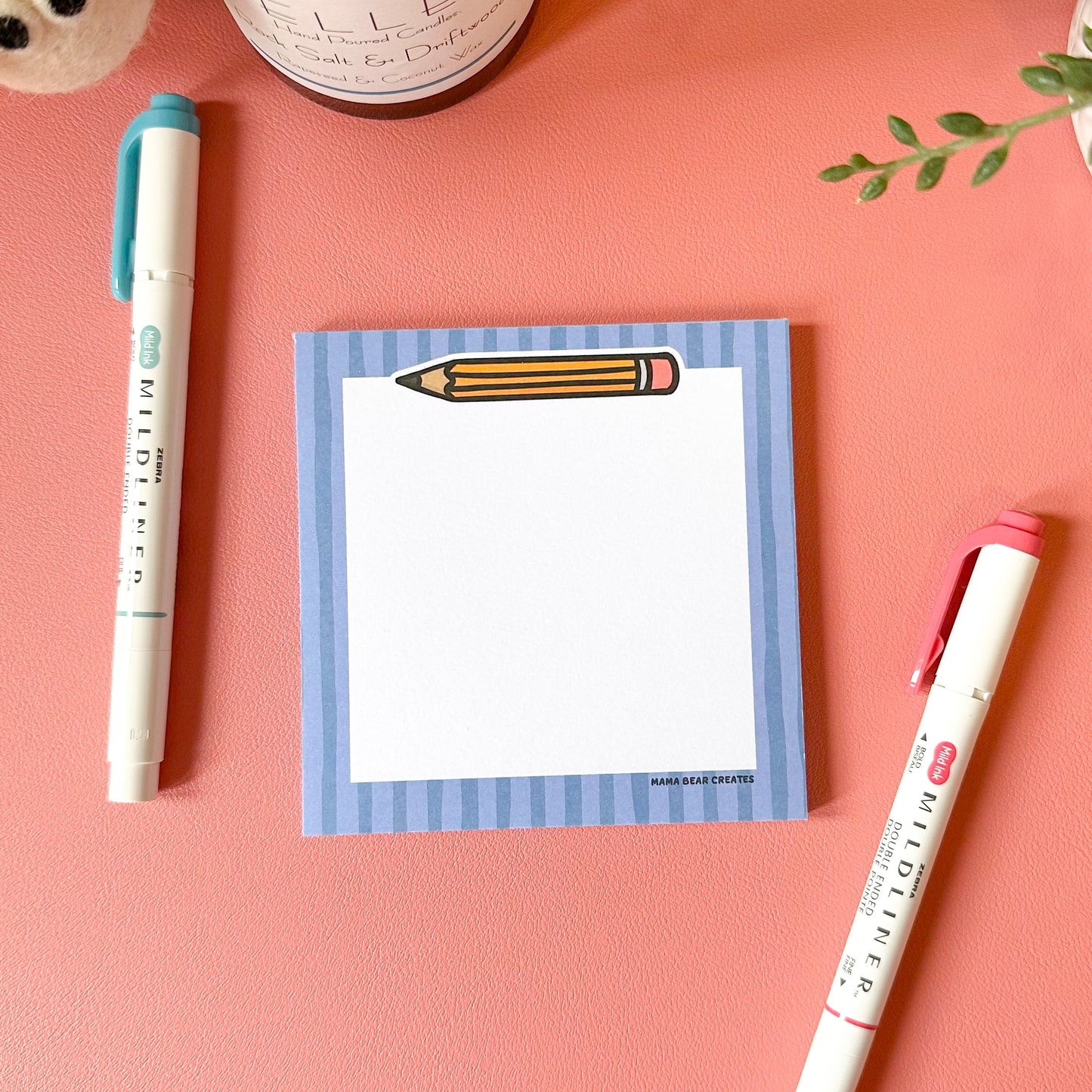 Notepad with a pencil design, pen, and candle on a pink surface