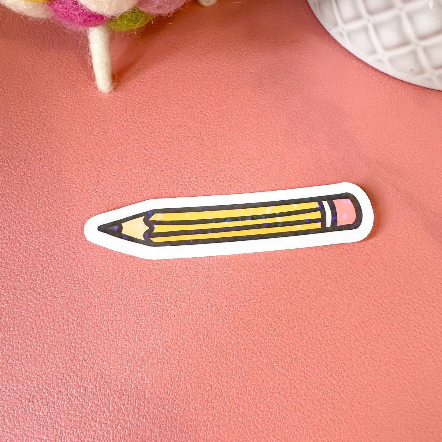 Sticker of a pencil on a pink surface with colorful pom-poms in the background
