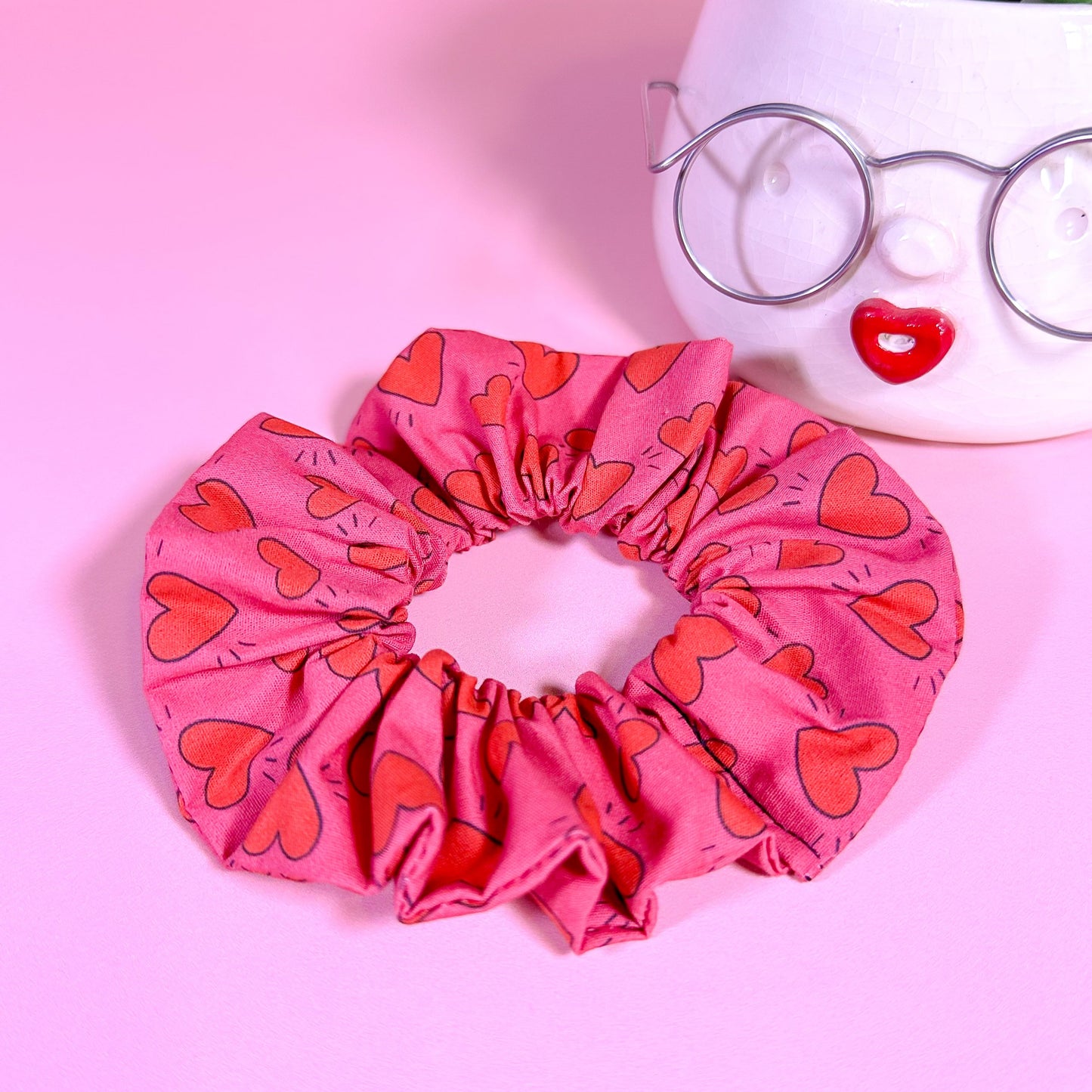 Pink scrunchie with heart pattern on a pink background with a plant and glasses.