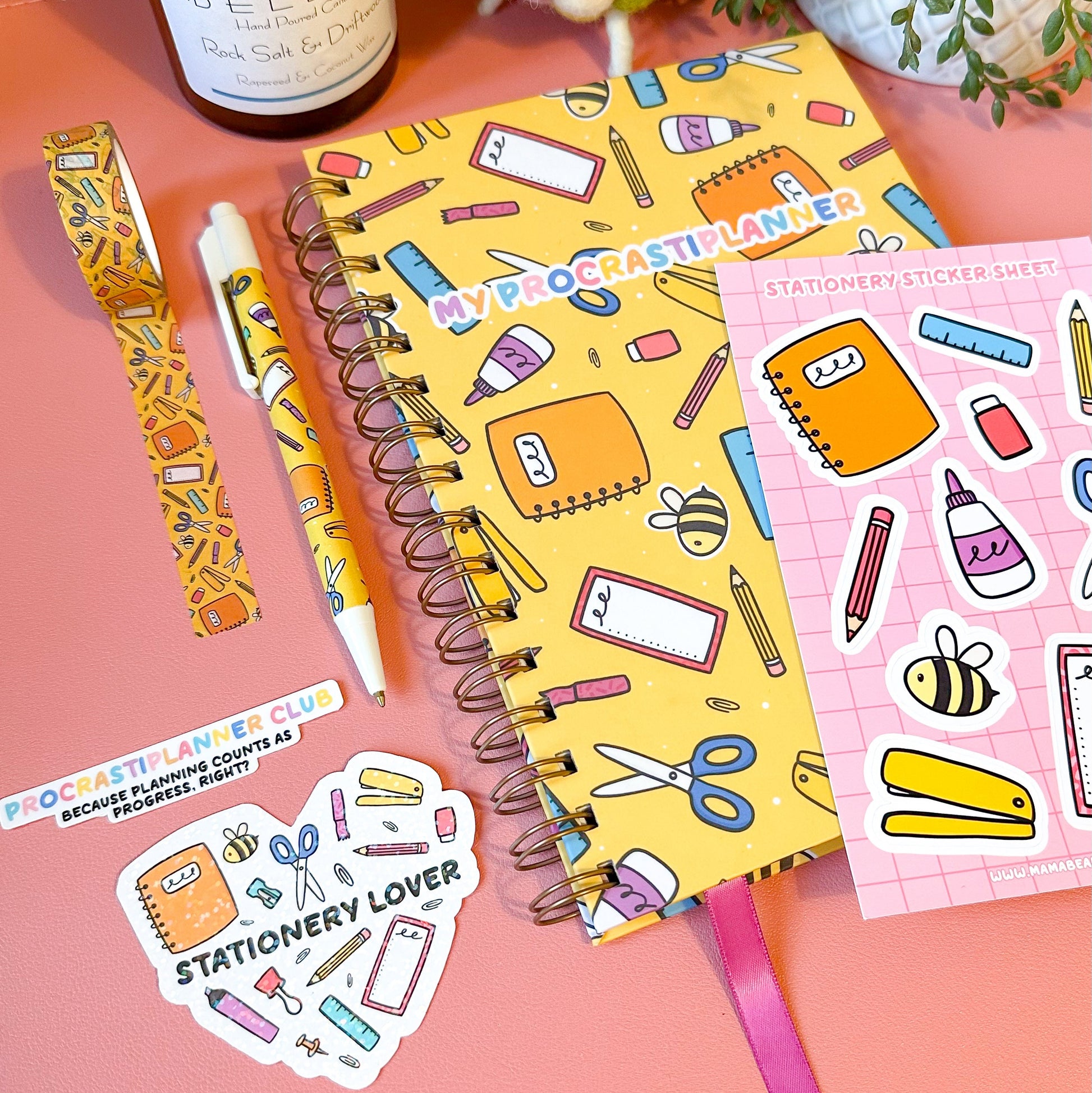 Stationery-themed items including a notebook, stickers, and a candle on a pink surface.
