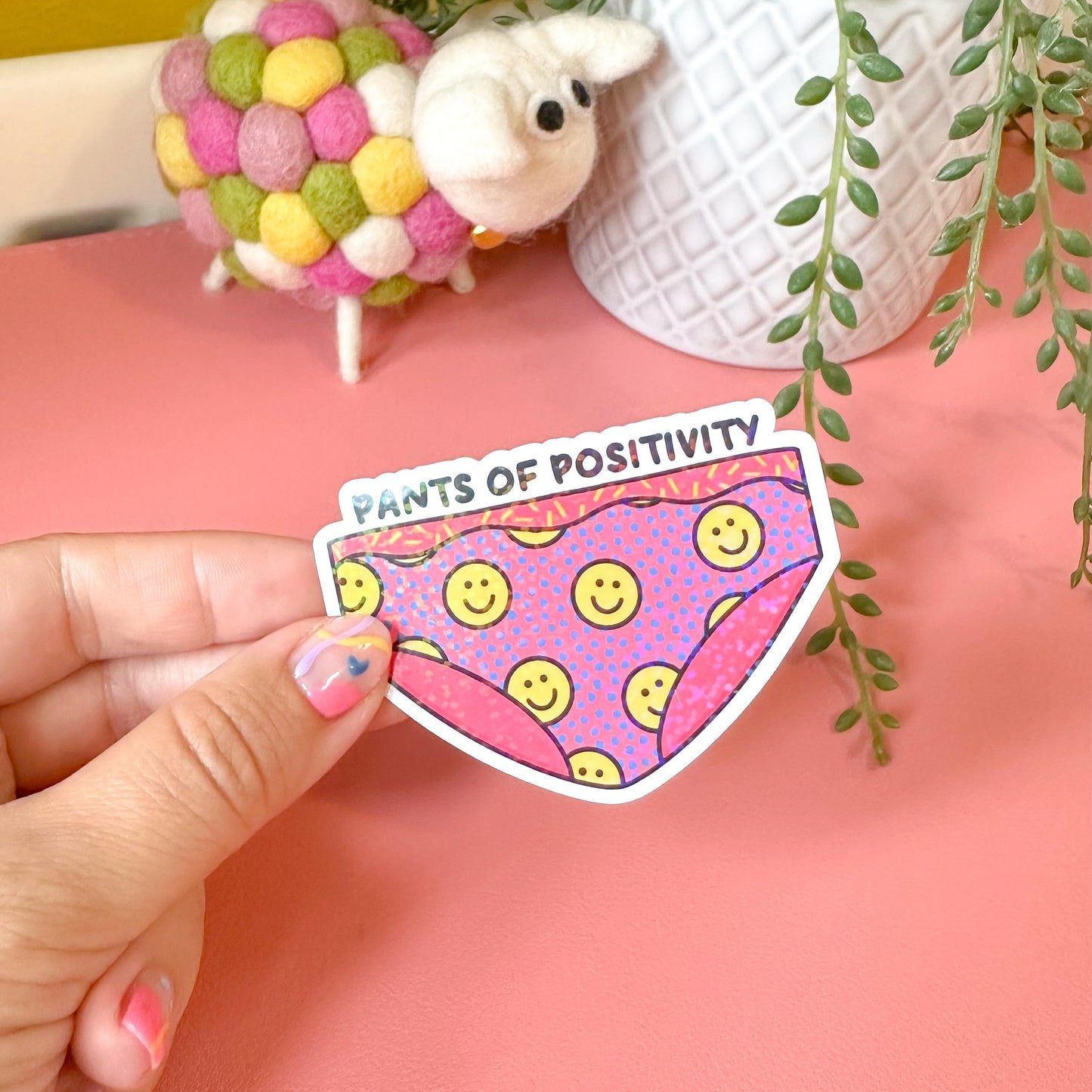 Hand holding a pink 'Pants of Positivity' sticker with smiley faces on a pink surface with decorative items.