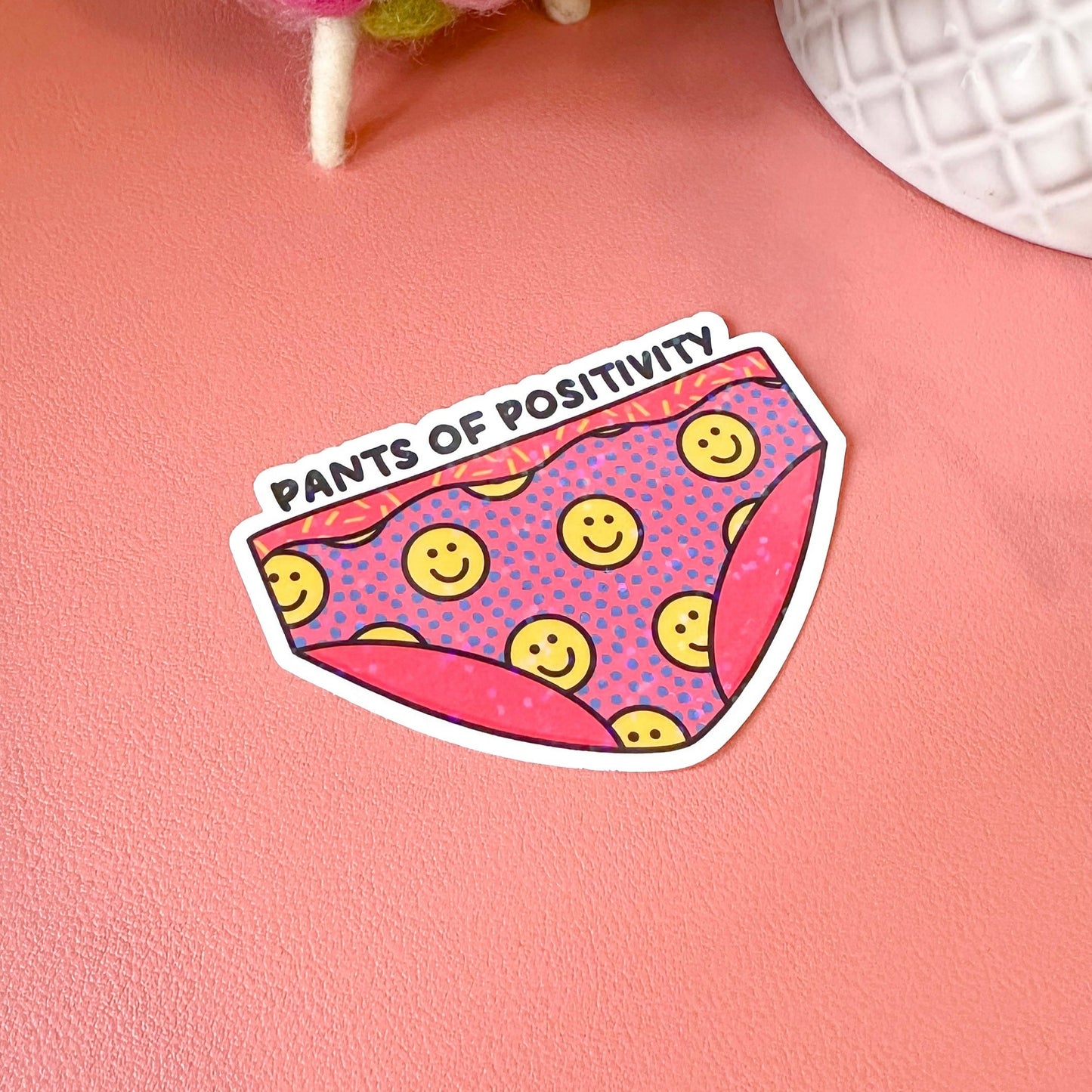 Sticker with 'Pants of Positivity' text and smiley face design on a pink surface.
