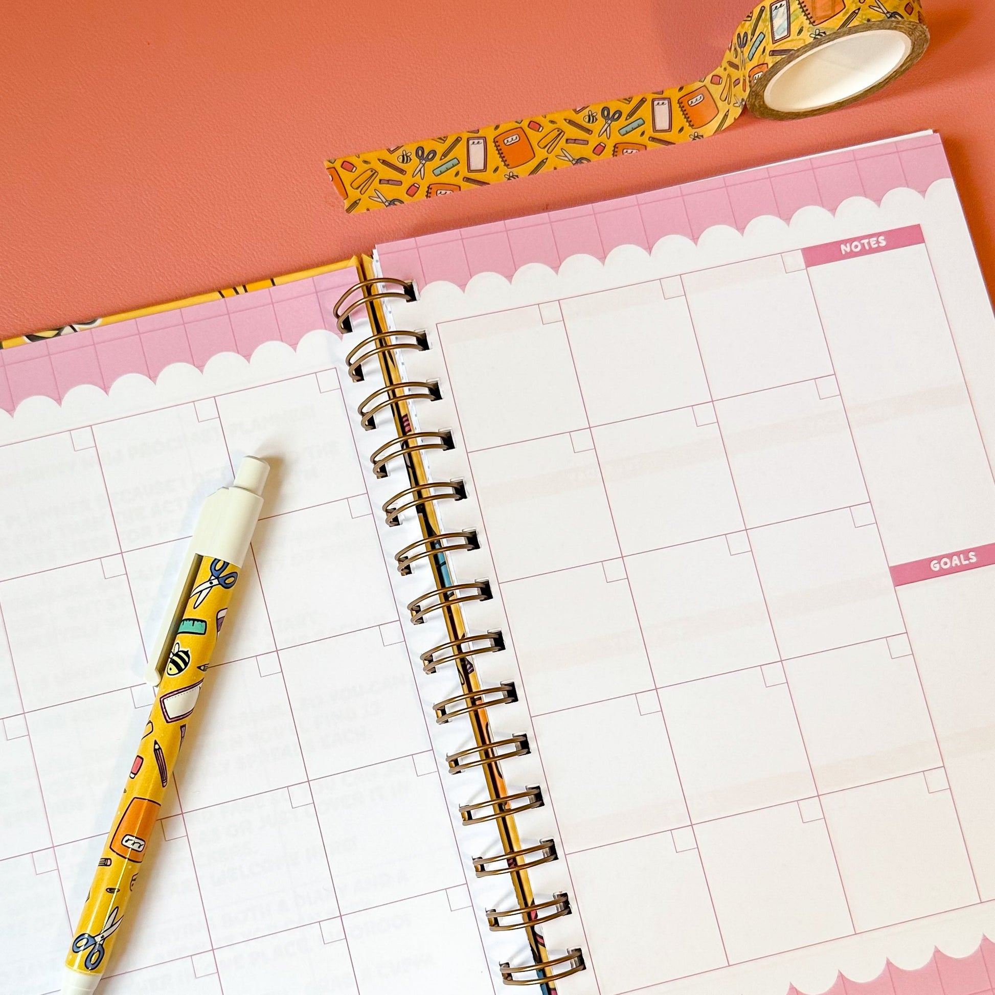 Spiral-bound planner with grid pages, a pen, and decorative tape on a pink background