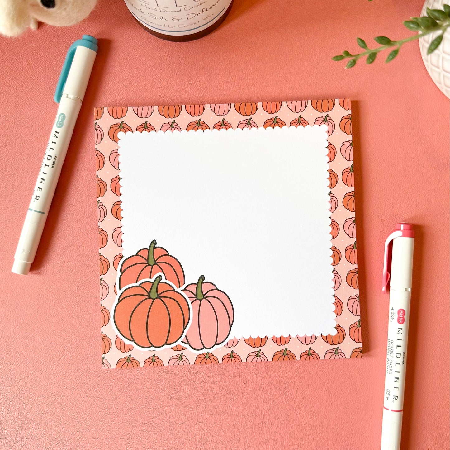 Notepad with pumpkin design on a pink surface with pens and a candle.