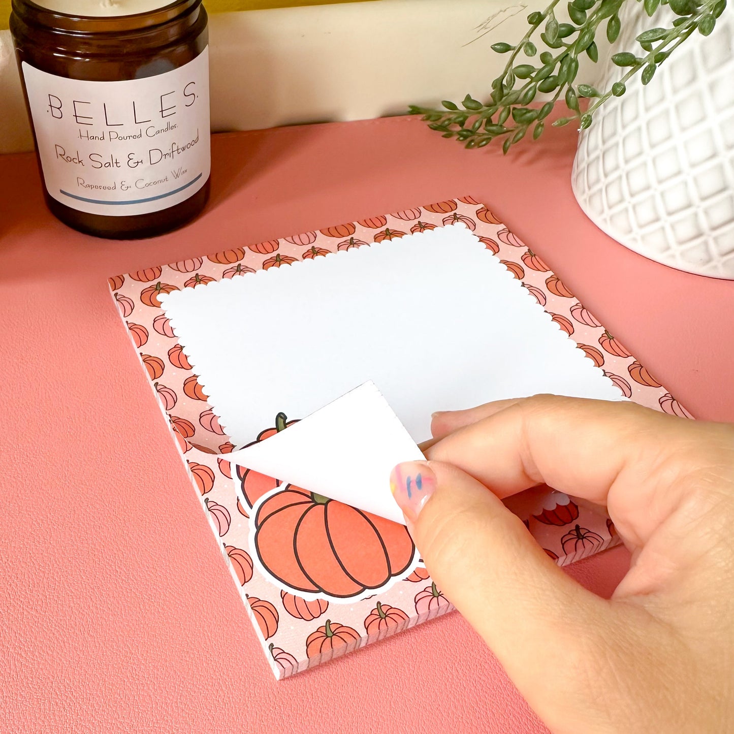 Hand holding a card with a pumpkin design on a pink surface next to a candle and plant.