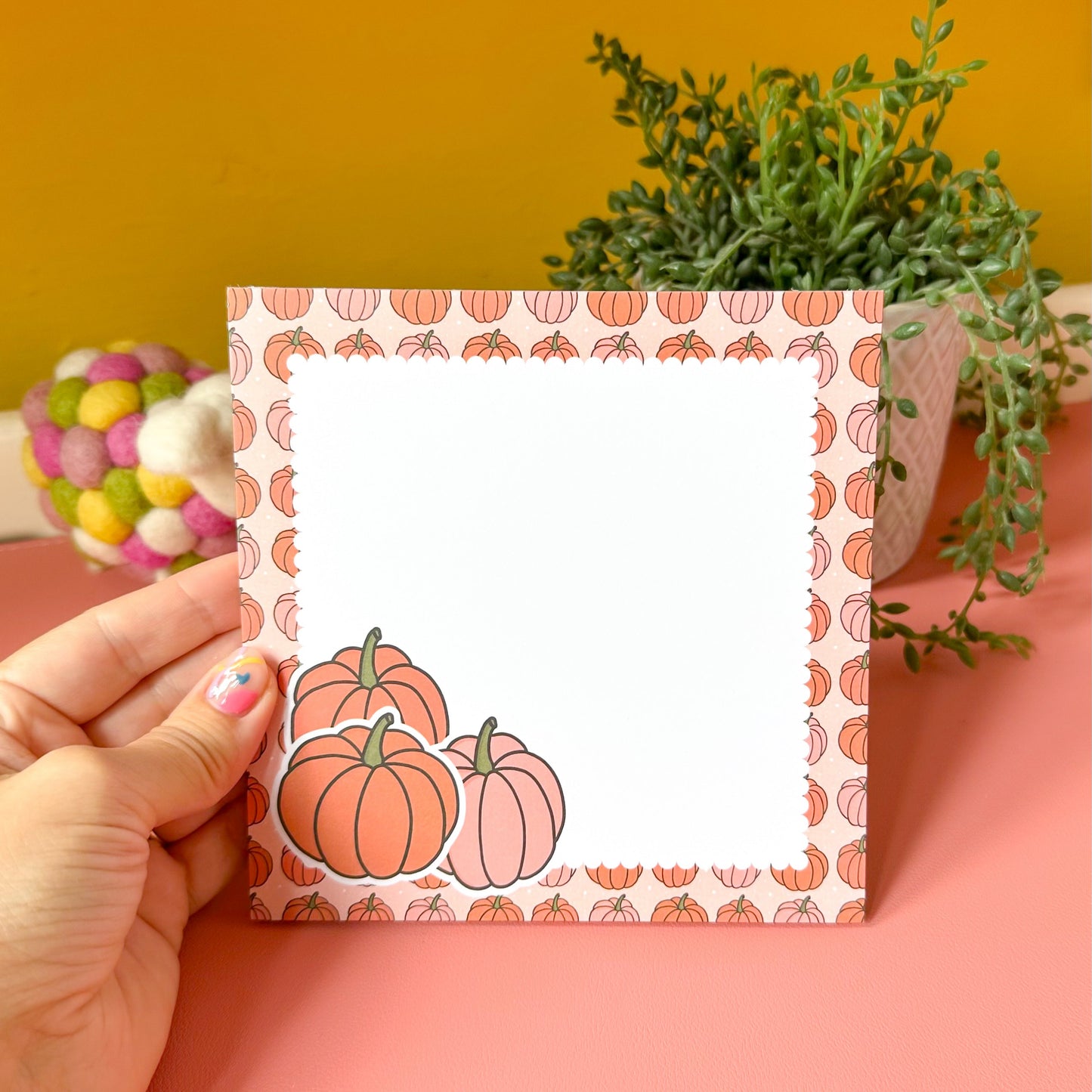 Hand holding a card with pumpkin design against a yellow wall with plants.