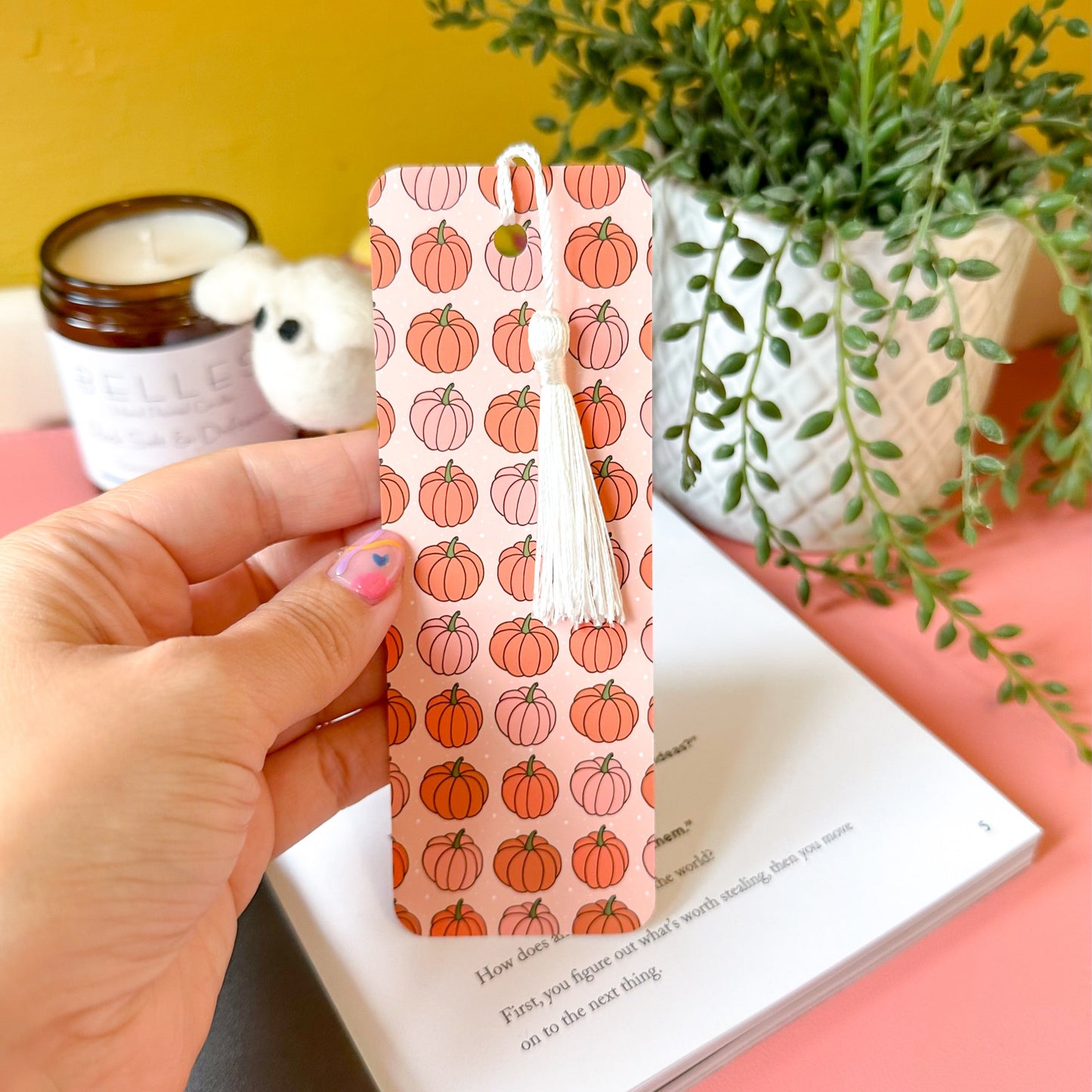 Hand holding a pink bookmark with pumpkin pattern on a pink surface with a plant and candle in the background.