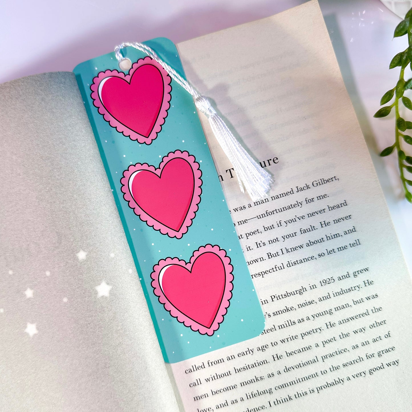A scalloped heart bookmark with three pink hearts on a blue background, placed inside an open book.