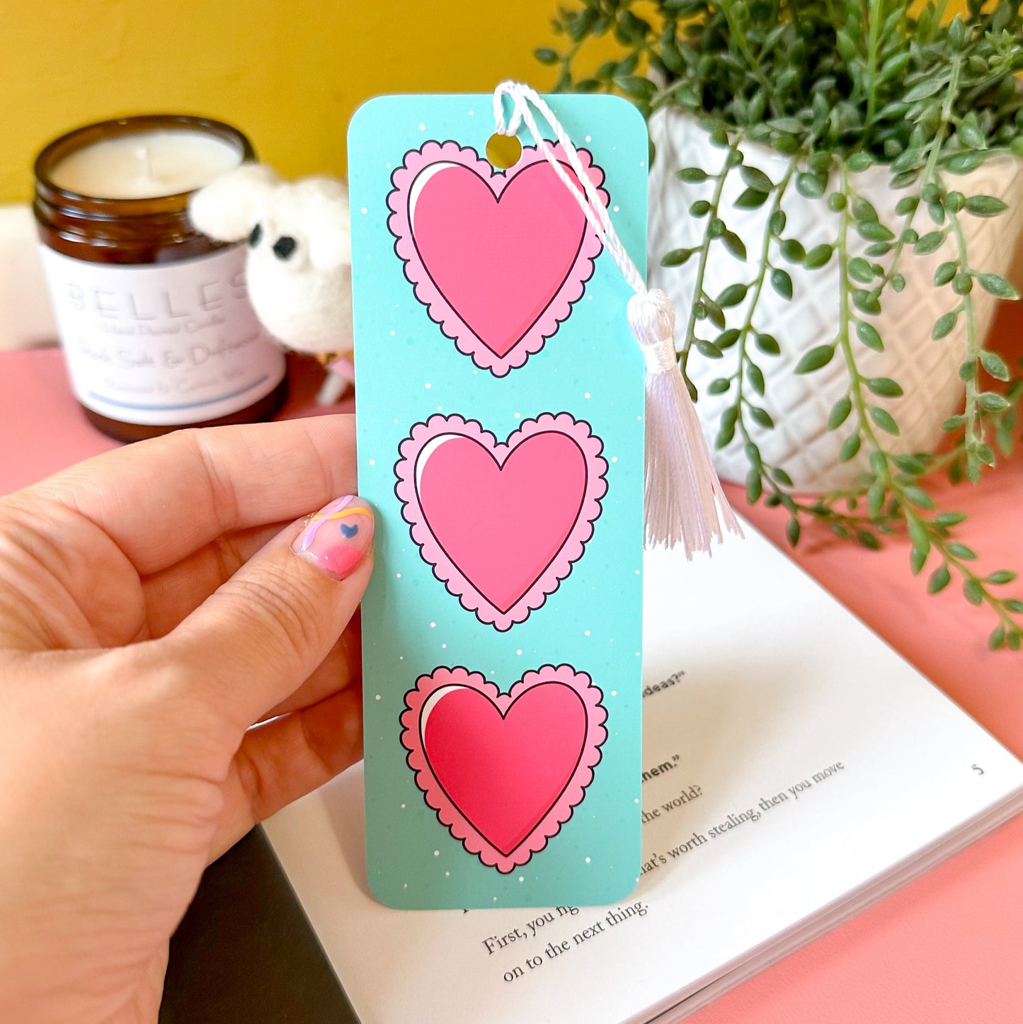 Bookmark with heart designs held by a hand on a book, with a plant and candle in the background.
