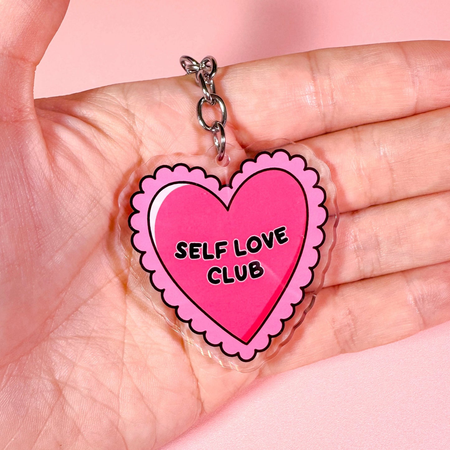 Heart-shaped 'Self Love Club' charm held in a hand against a pink background