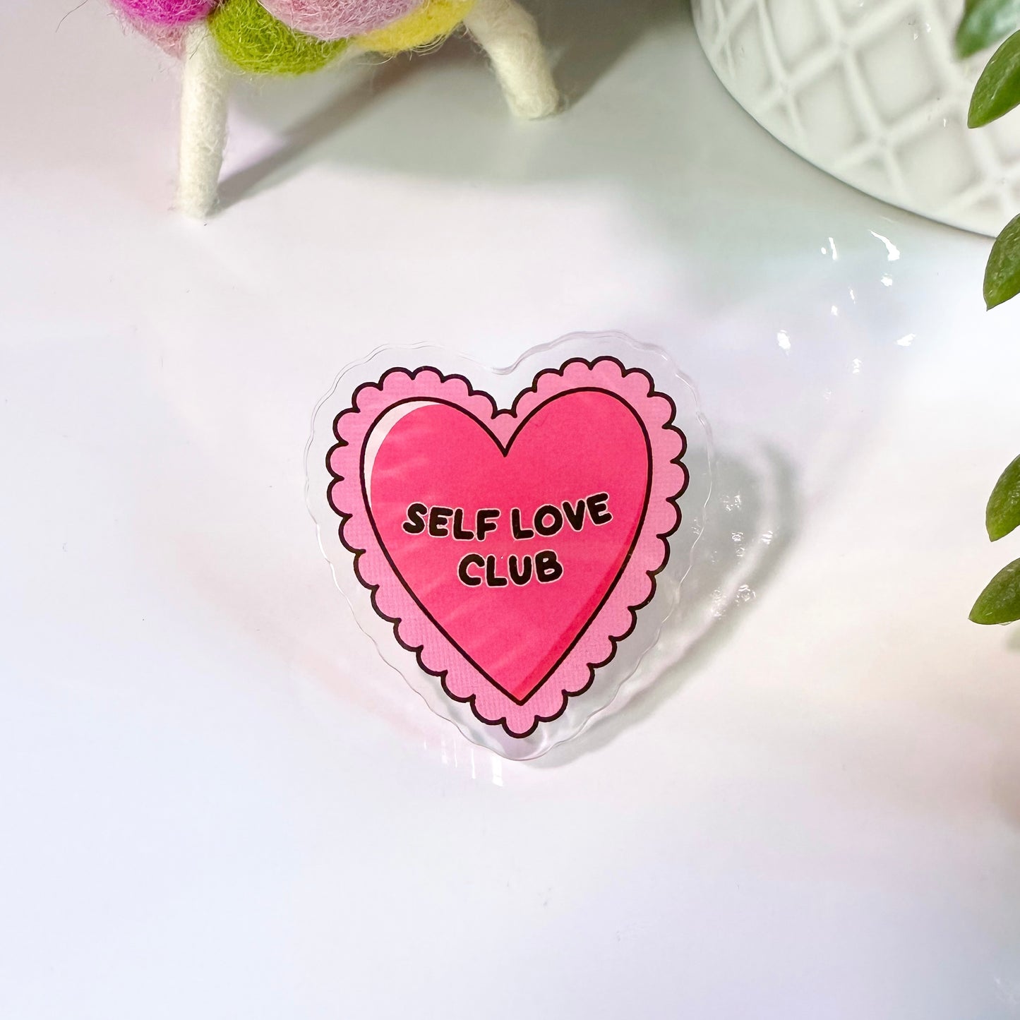 Acrylic pin badge with a pink heart shape, black text reading 'SELF LOVE CLUB', and scalloped pink edges on the badge, displayed on a white background.