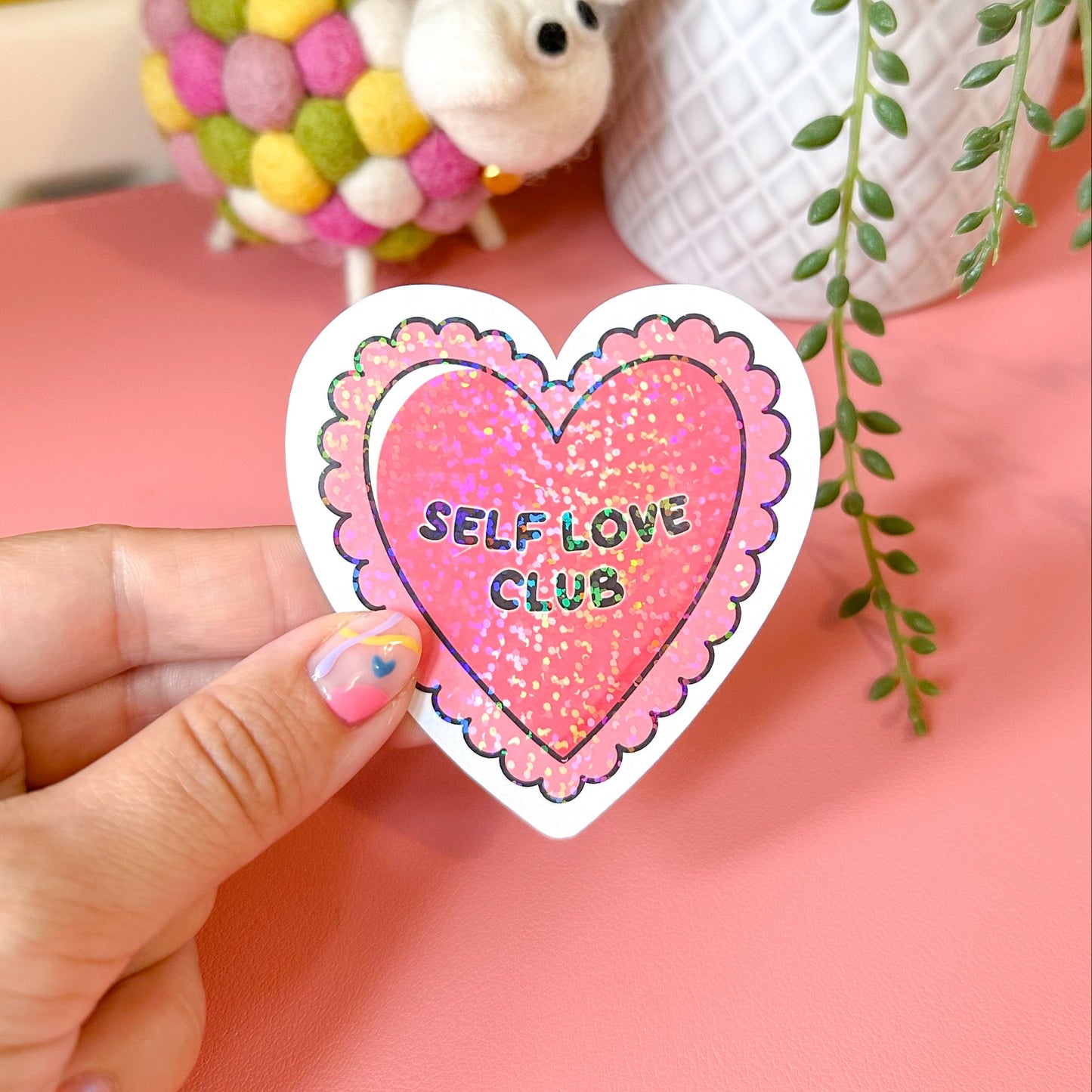 Heart-shaped 'Self Love Club' sticker held by a hand with a pink background