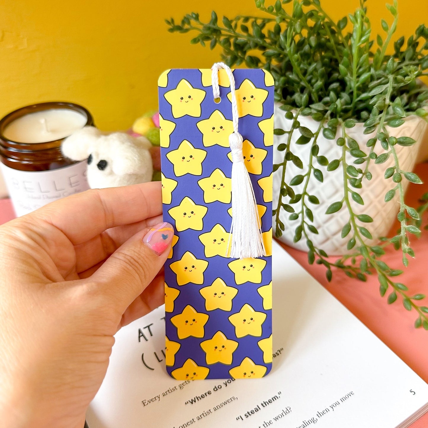 Hand holding a yellow star-patterned bookmark with a plant and candle in the background