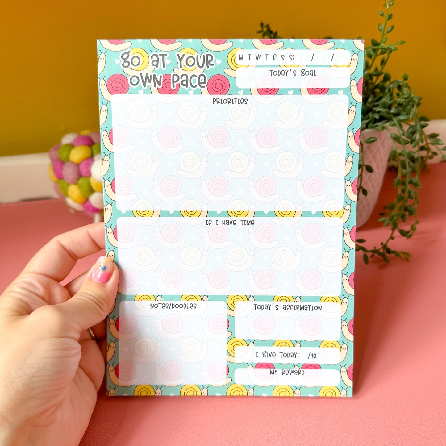 Hand holding a colorful notepad with a yellow wall and pink surface in the background