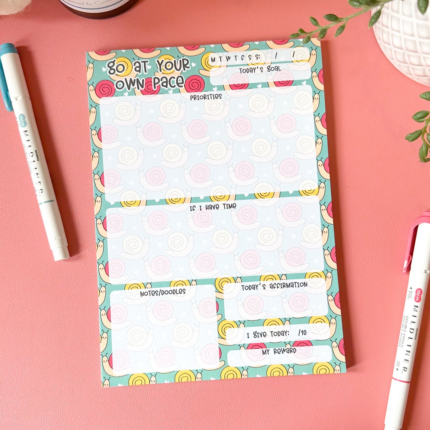 Colorful notepad with a decorative border on a pink surface with pens and a plant.