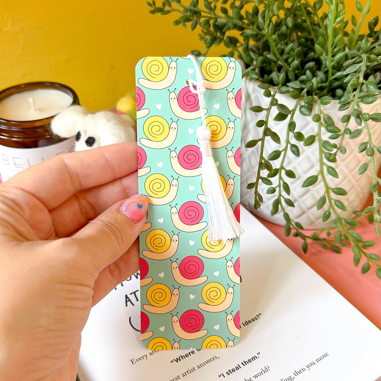 bookmark with snail pattern held by a hand, yellow wall and plant in the background