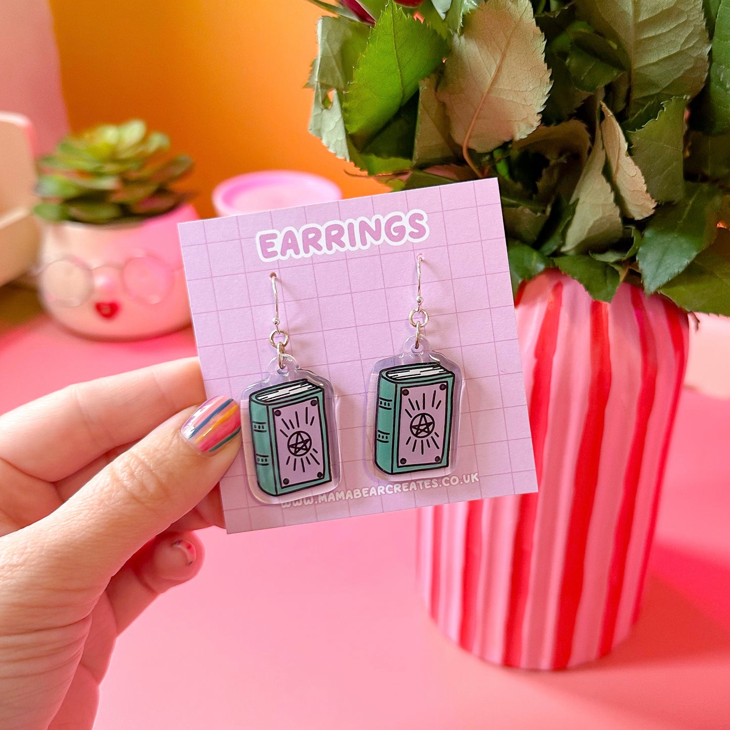 A pair of acrylic earrings designed to resemble spell books, held up against a pink background with a plant and a cup.