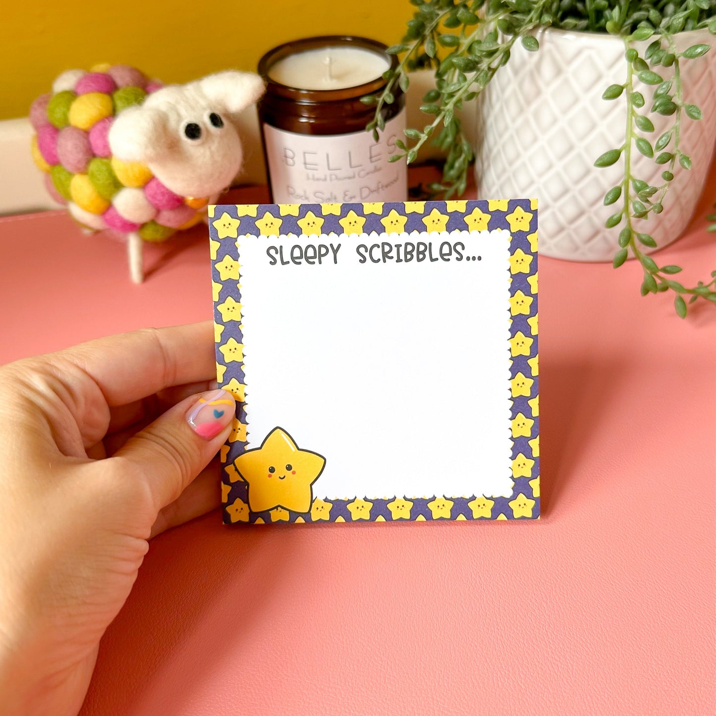 Hand holding a notepad with star design and 'Sleepy Scribbles' text on a pink surface with decorative items.